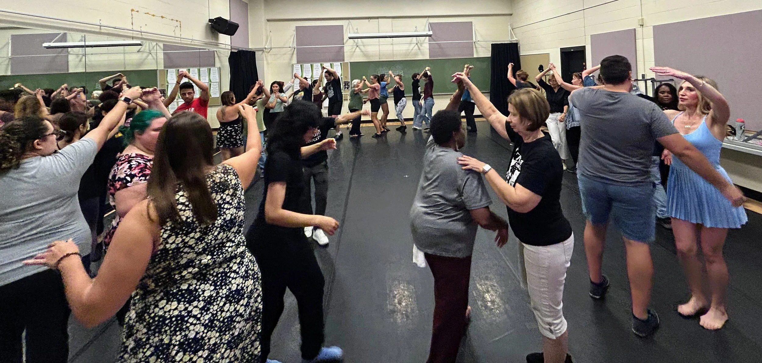 A diverse group of people dancing together in a dance studio with mirrors and a black floor, forming circles and pairs.