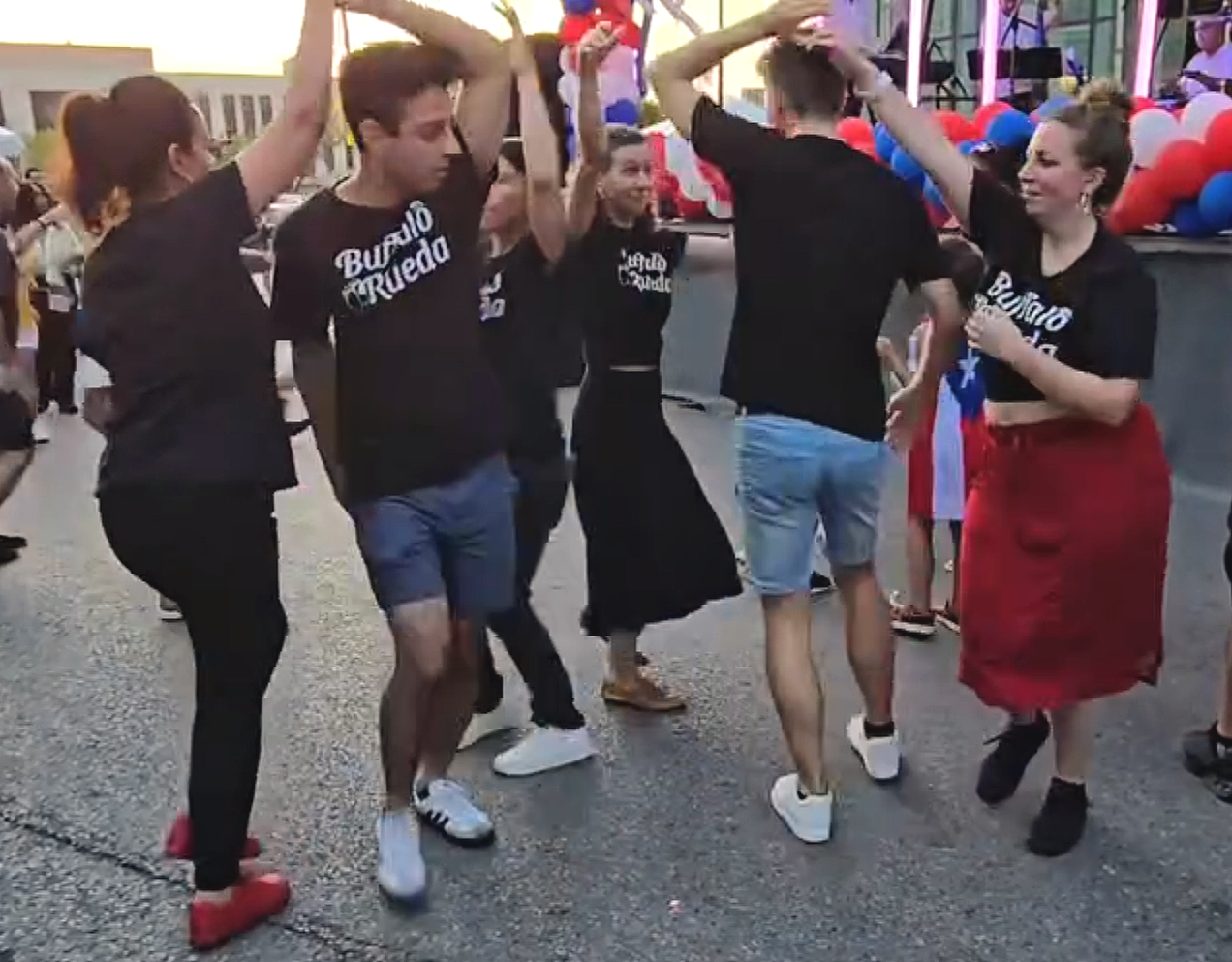 Group of people dancing together at an outdoor event decorated with red, white, and blue balloons, with some wearing matching black t-shirts, celebrating at sunset.