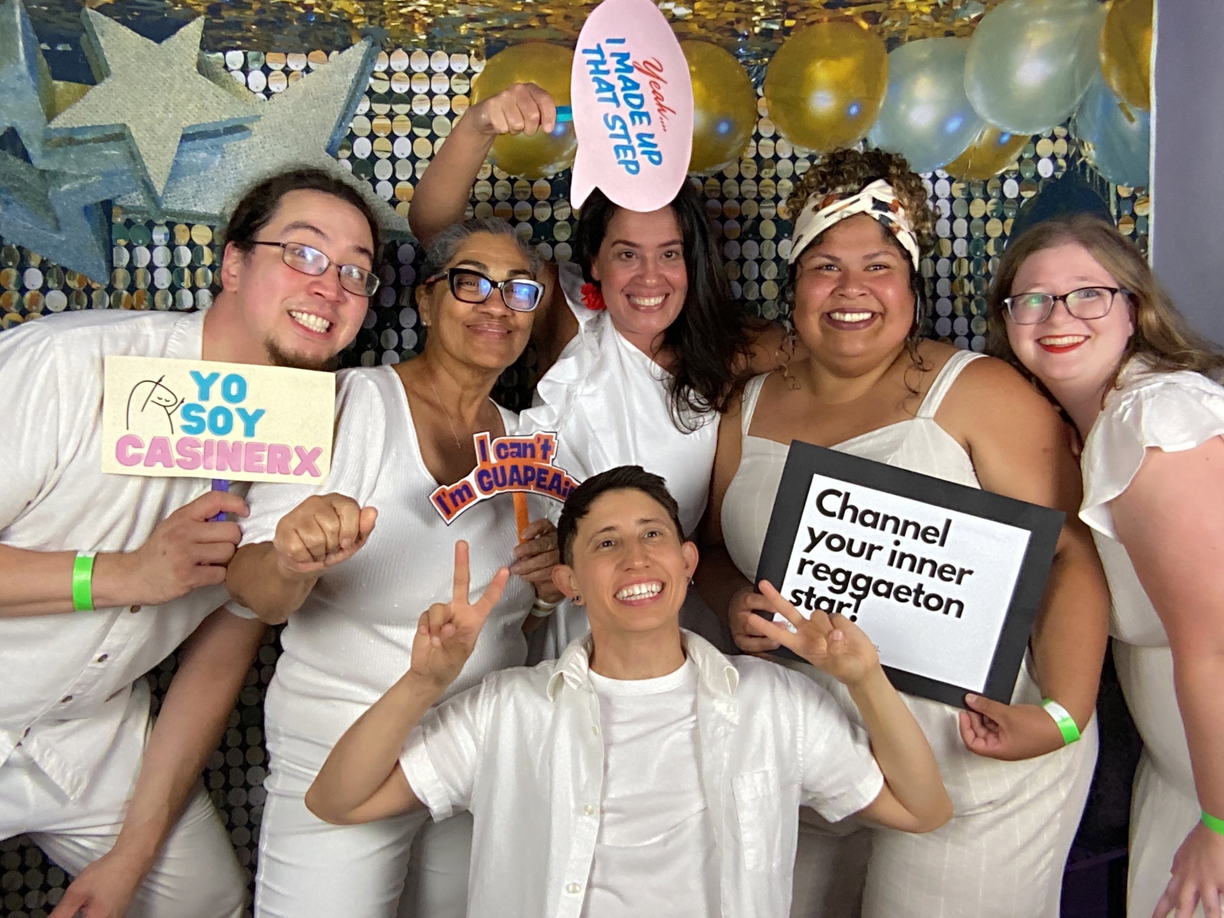 Group of six women and one man holding signs and smiling at a celebration. The background is decorated with gold and silver balloons and stars.