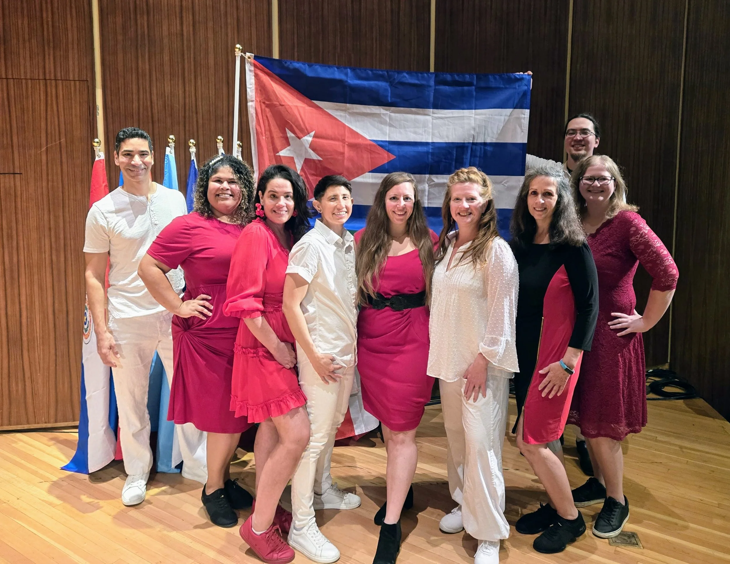 A group of ten diverse people standing in front of a Cuban flag and other flags, smiling and posing for a photo in a wood-paneled room.