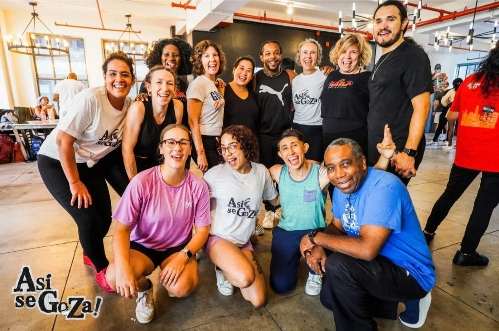 Group of diverse smiling people in casual clothing posing indoors, with some kneeling and some standing, in a lively and cheerful setting.