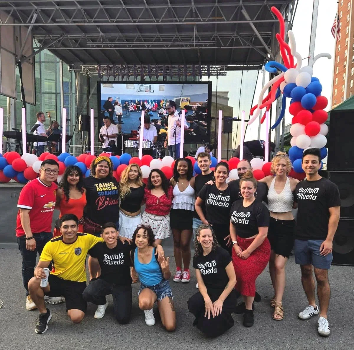 Group of people smiling and posing for a photo at an outdoor event, with a stage decorated with red, white, and blue balloons, and a large screen showing a live view of the stage. The team wears coordinated shirts, some with "Buffalo Rueda" printed on them.