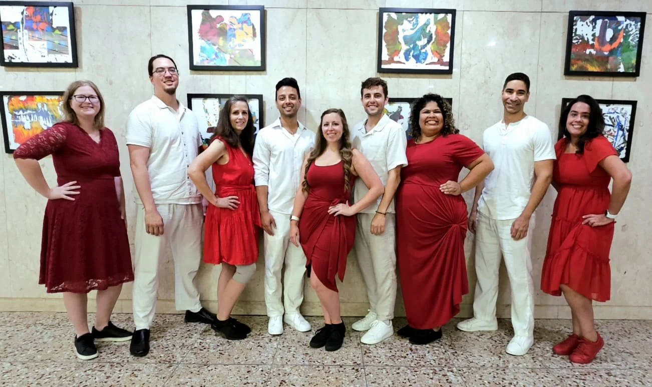 Group of ten people standing in front of an art display on a light-colored wall, all smiling and dressed in shades of red and white.