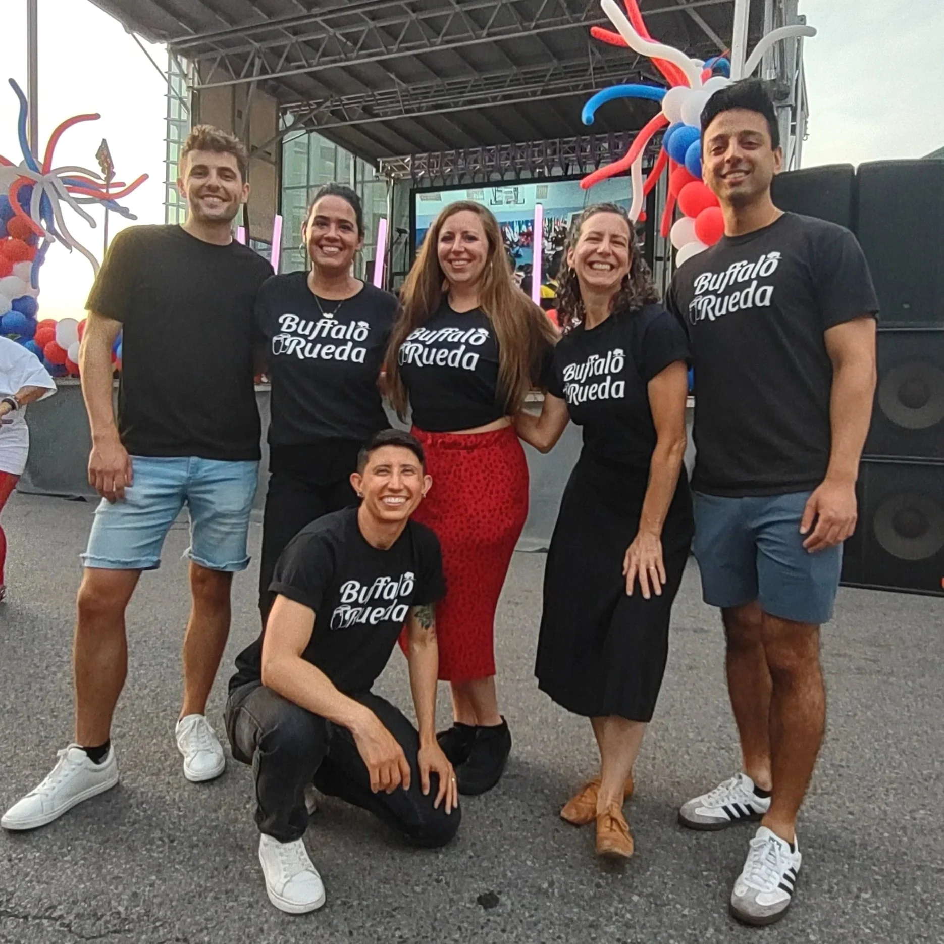 Group of six people smiling at an outdoor event, wearing matching black t-shirts with "Buffalo Rueda" printed on them, standing in front of a stage decorated with red, white, and blue balloons and colorful lights.