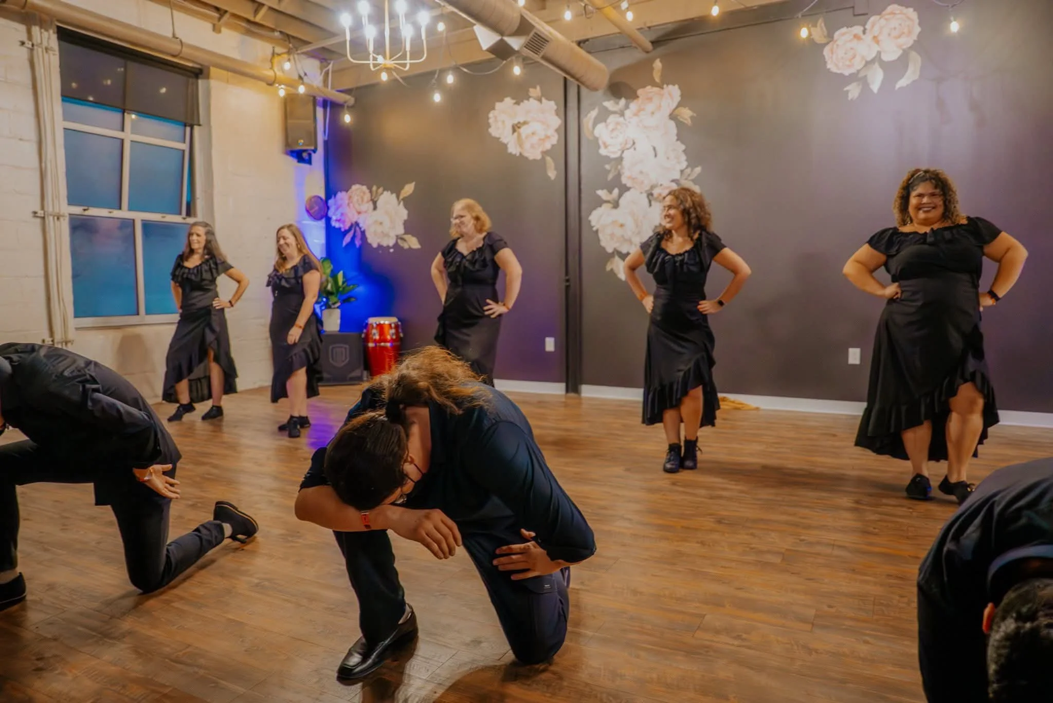 Women dressed in black dresses dancing on a wooden-floor stage with floral murals on dark wall, string lights, and window with blinds, with some people kneeling in front.