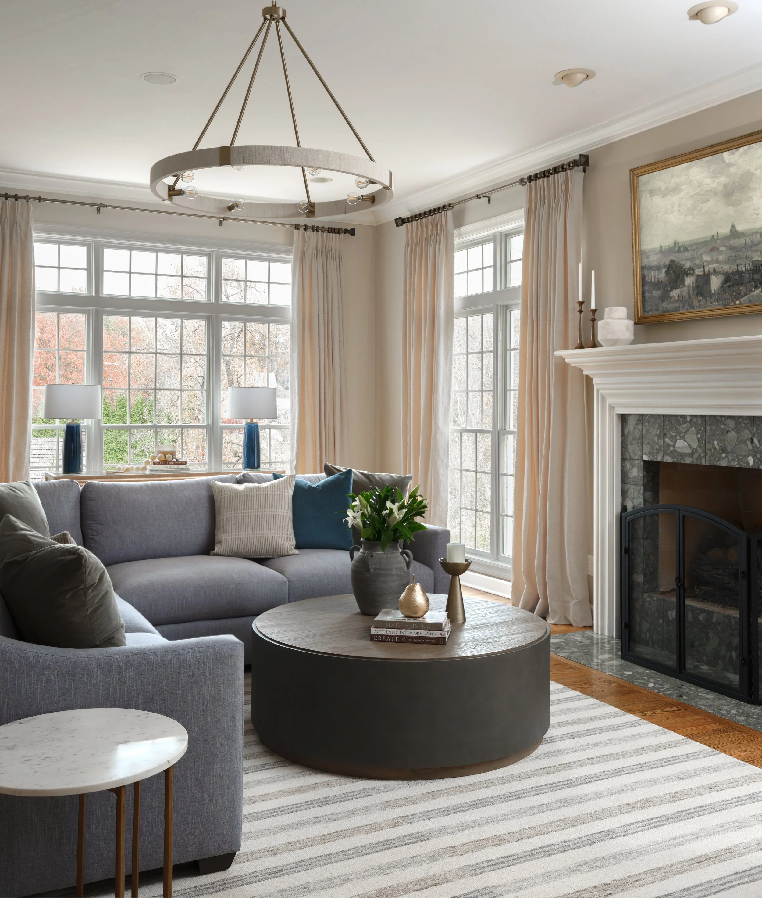 Living room with large windows, cream curtains, blue sofa with pillows, round coffee table with decorative items, fireplace with mantle, hanging ceiling light, and a framed tv.