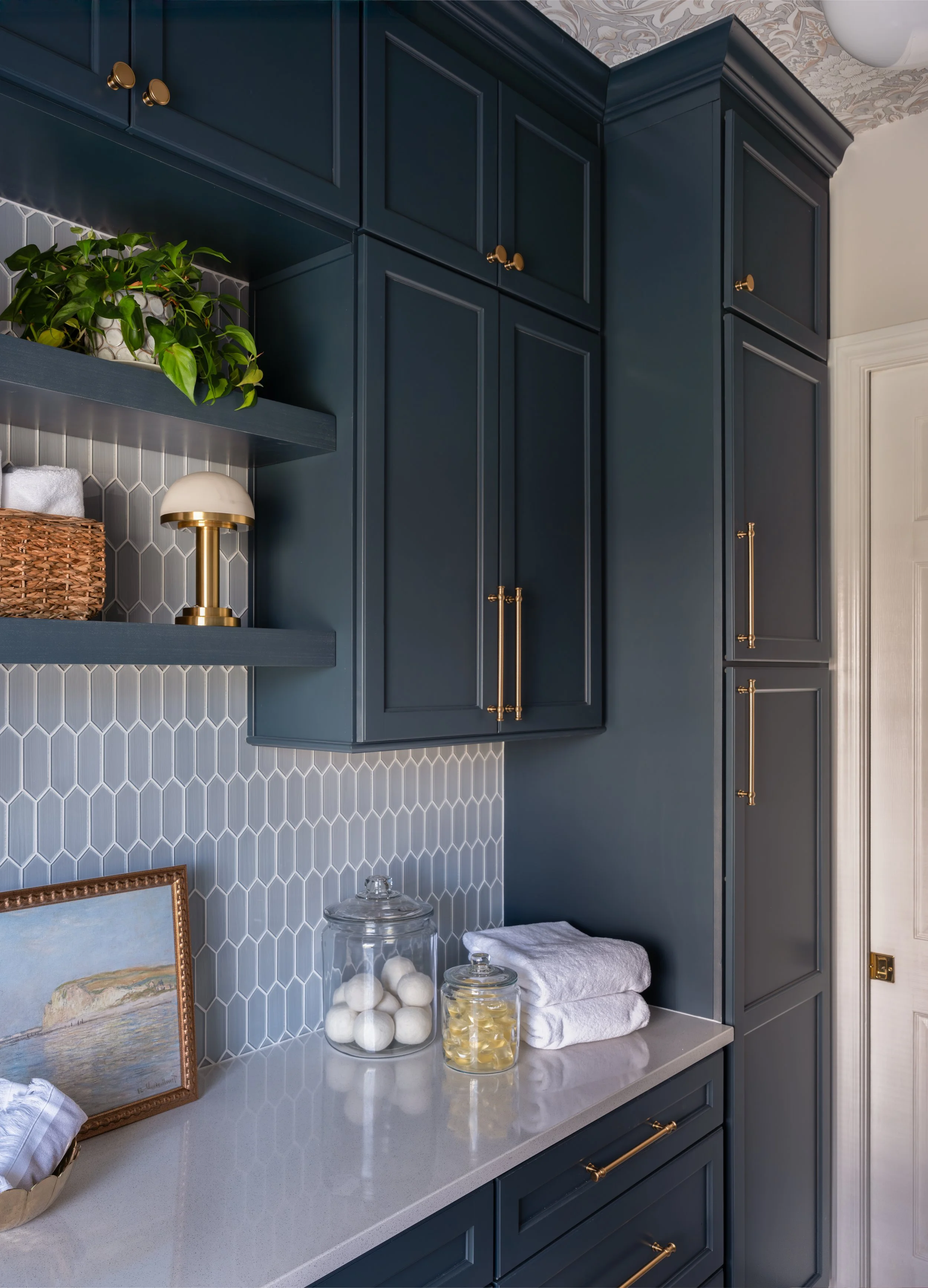 A corner of a modern kitchen with navy blue cabinets, gold hardware, and a white countertop. There are decorative jars, a towel, a framed picture, and a small lamp on the countertop. A plant is on a shelf above the counter.
