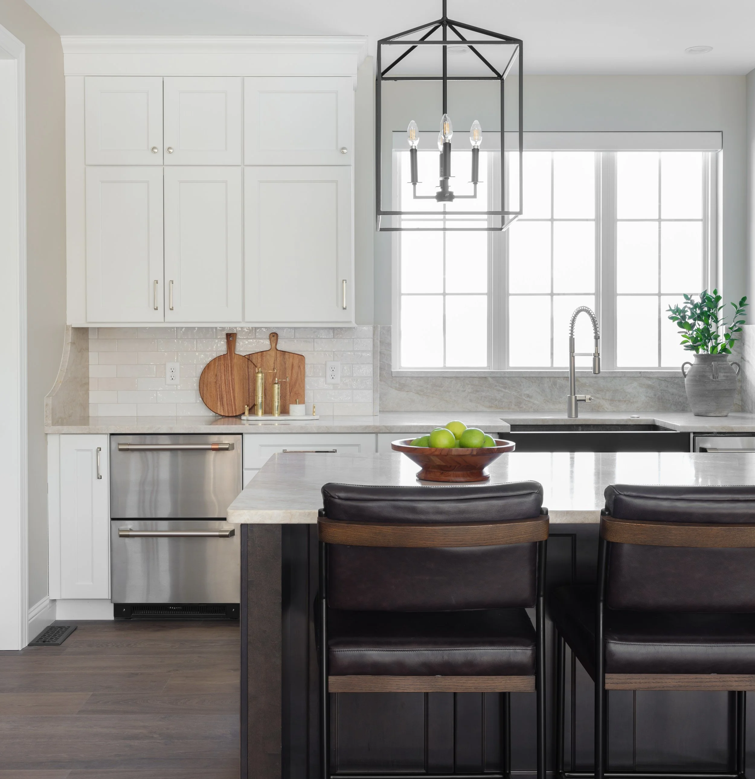 Modern kitchen with white cabinets, gray countertops, large window, black and wood chairs at a kitchen island, bowl of green apples, and decorative cutting boards on the wall.