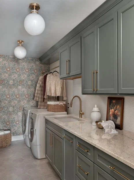 Laundry room with green cabinets, light countertop, washer and dryer, floral wallpaper, hanging clothes, basket, and decorative items.