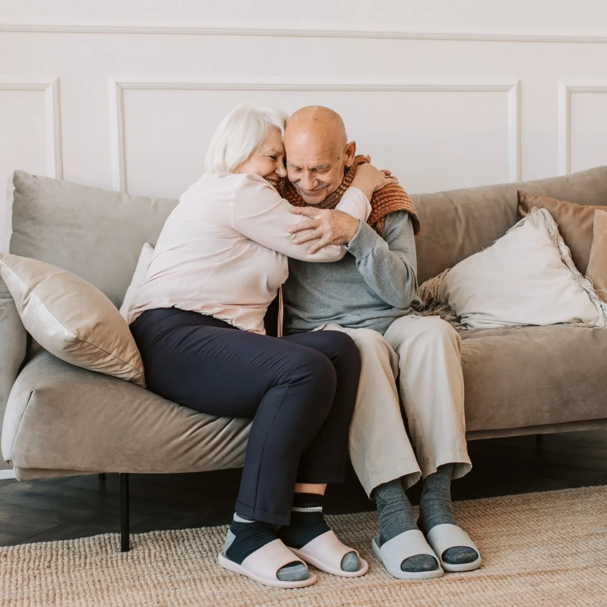An elderly woman and man sharing a joyful hug on a sofa in a cozy living room.