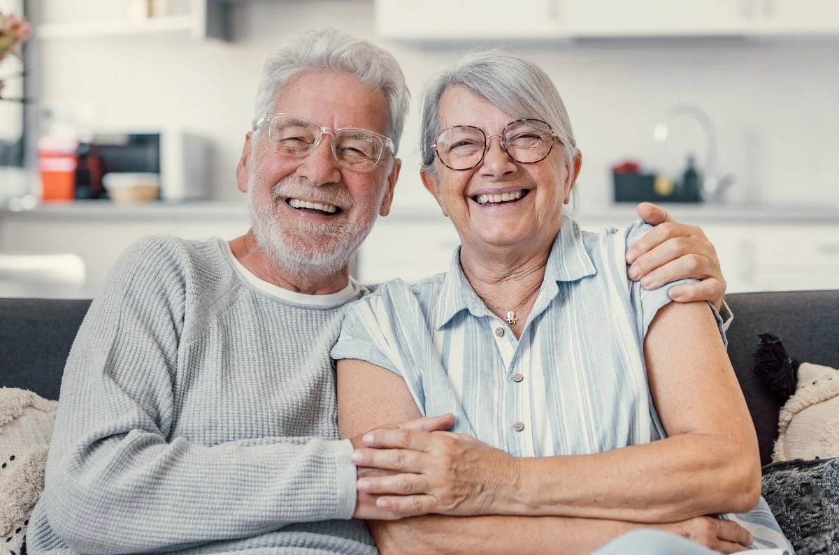 An elderly couple sitting on a couch, smiling and hugging each other in a cozy, modern kitchen.