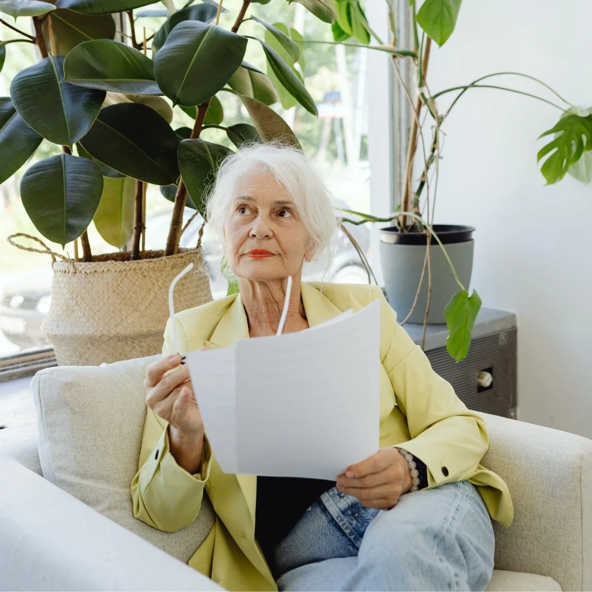 An elderly woman with white hair sitting on a cream-colored sofa in a bright room, holding a piece of paper and a pair of glasses, surrounded by large green houseplants.