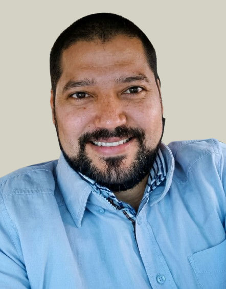 Portrait of a man with short dark hair and a beard, smiling, wearing a light blue collared shirt, against a plain light background.