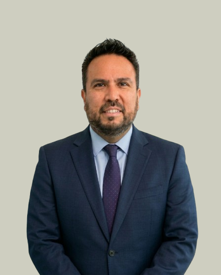 Professional man in a dark blue suit and tie, smiling, standing against a plain light background.