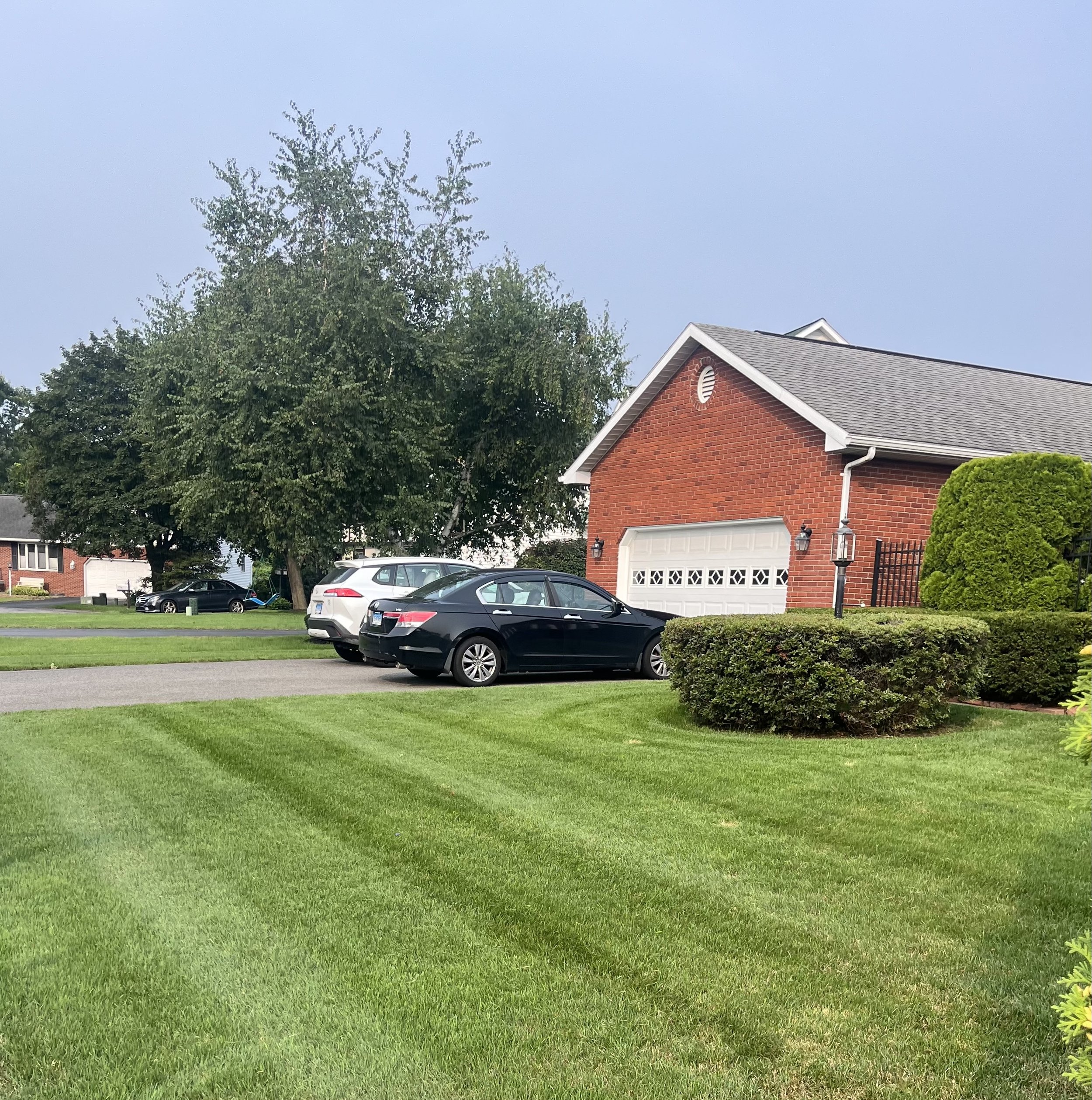 A suburban residential scene showing a well-maintained lawn, a driveway with parked cars, and a red brick garage with a gray door. There are trees and bushes around the property and neighboring houses visible in the background under a cloudy sky.