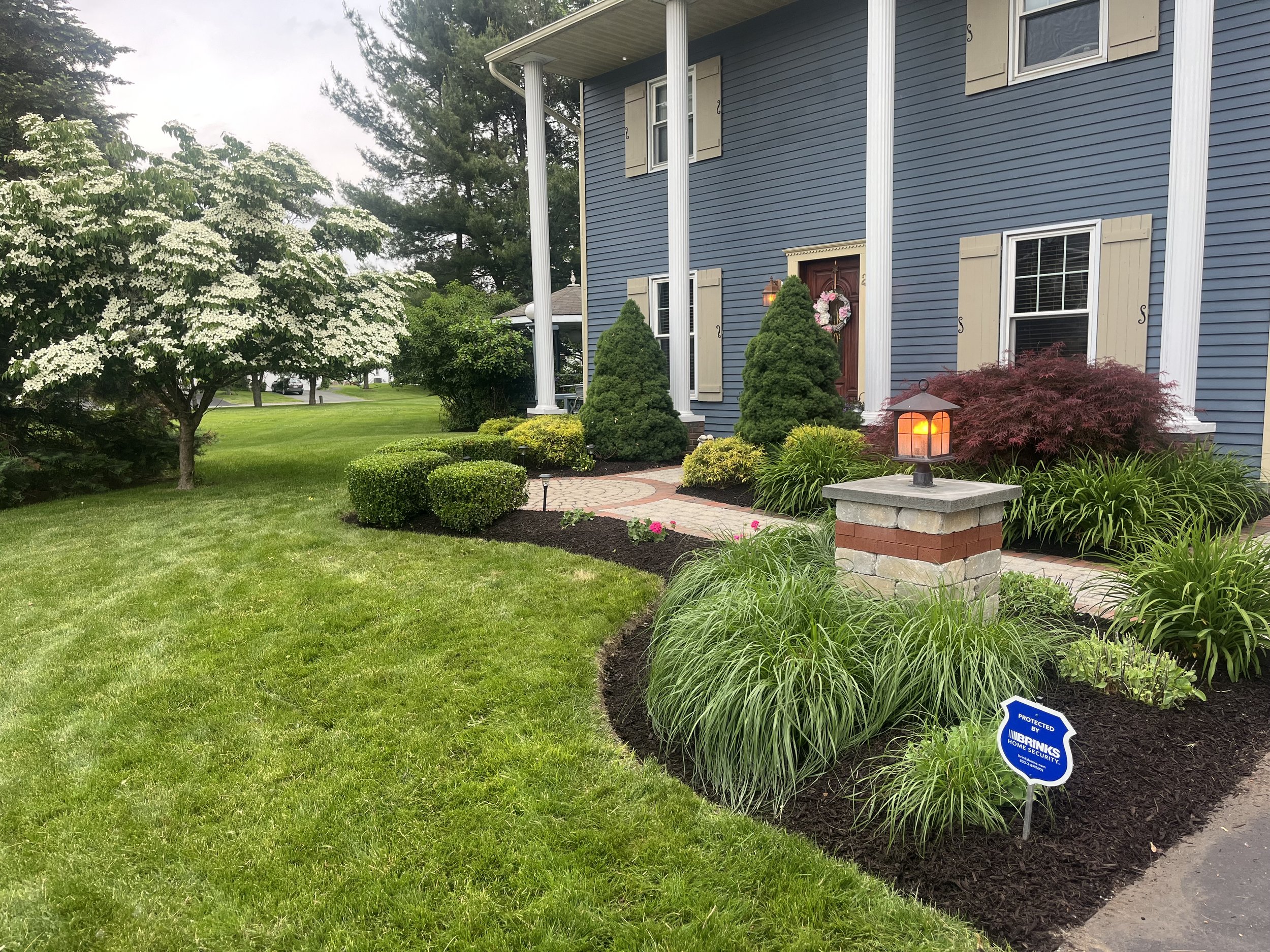 Front yard of a blue house with beige shutters, manicured garden with small shrubs, ornamental trees, and a brick pathway, security sign, and a lantern on a stone pillar near the porch.