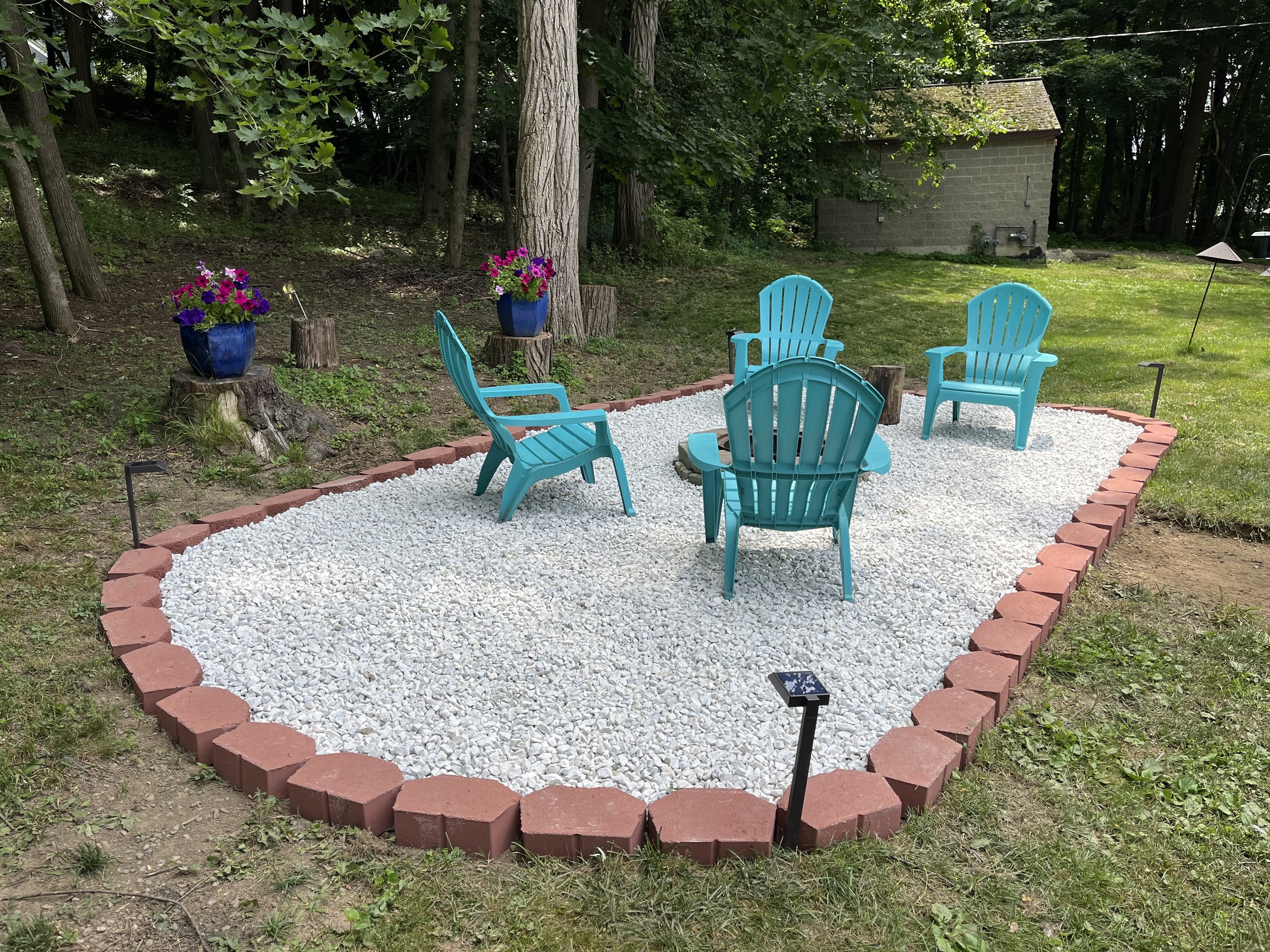Small garden area with white gravel ground bordered by red bricks, surrounded by trees and grass. There are five blue Adirondack chairs arranged in a circle and two large blue flower pots with pink and purple flowers on tree stumps.