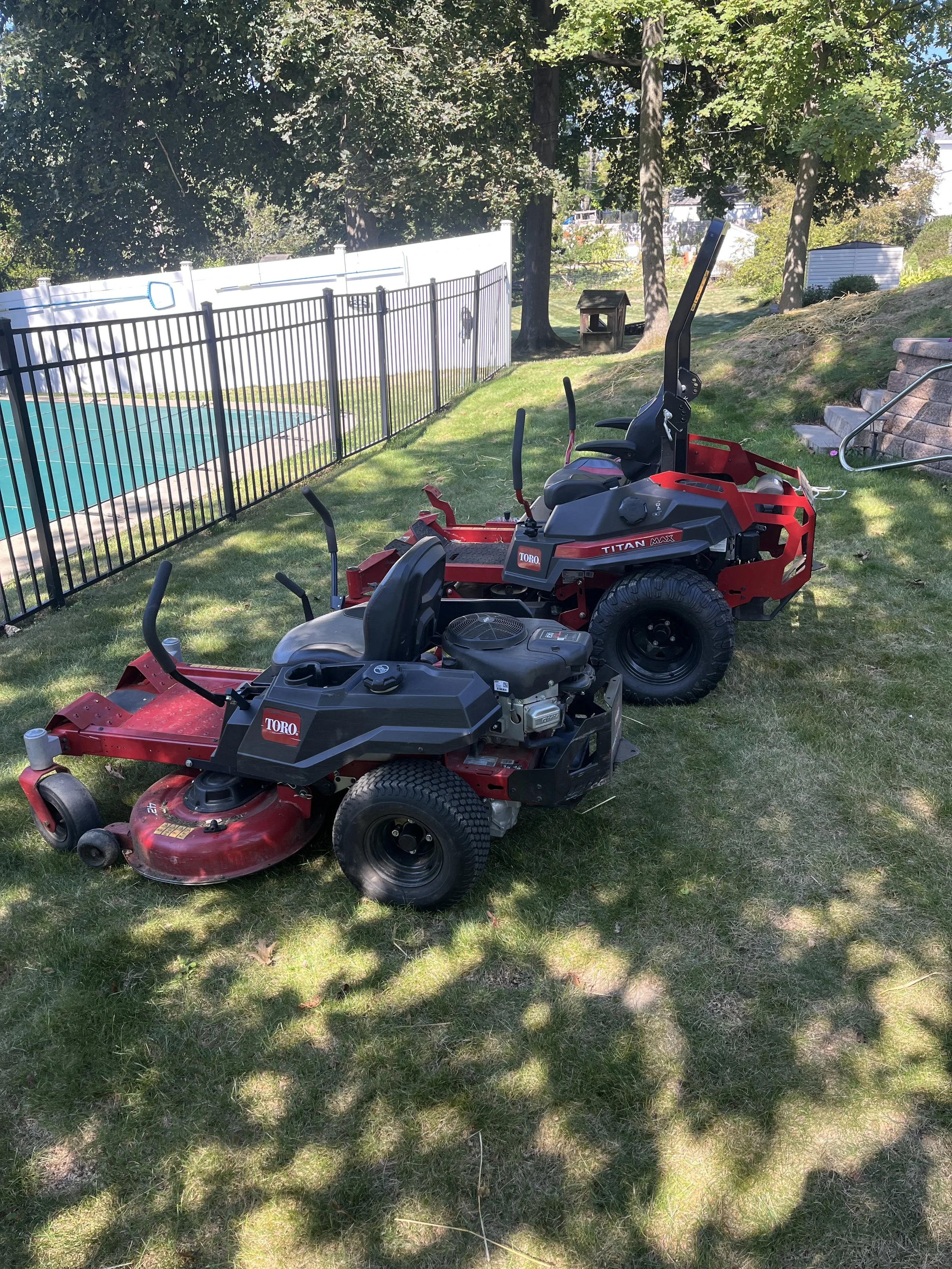 Two Toro brand lawnmowers parked on a grassy backyard with trees, a pool fence, and a dog house in the background.