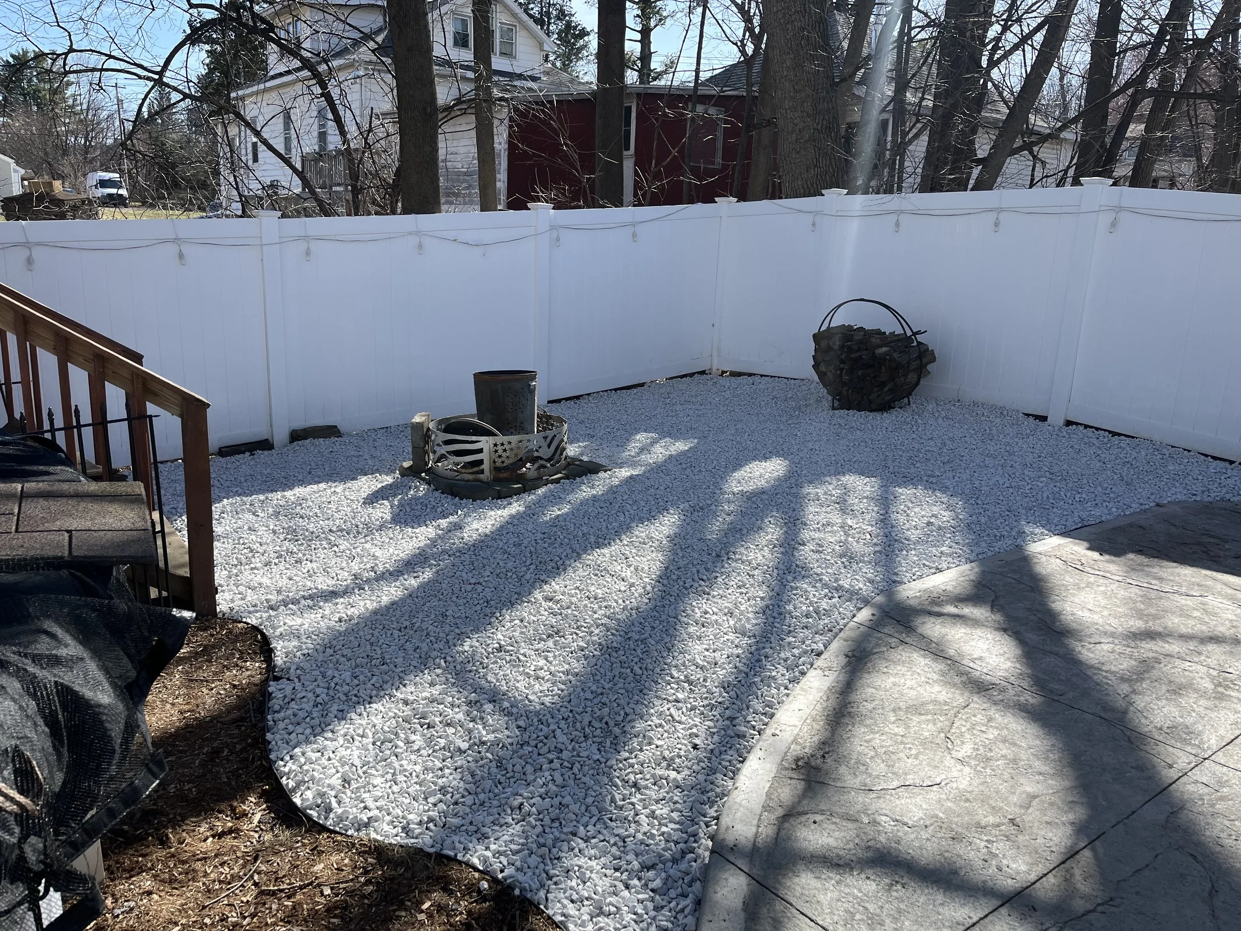 Backyard with white gravel ground, white privacy fence, trees, and a house in the background. There are firewood logs in a metal holder and a small decorative fire pit with a bucket on top. Part of a concrete patio and a wooden staircase are visible.