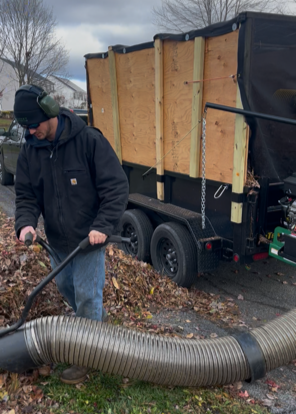 A person wearing a black jacket, gloves, and a cap using a large vacuum or blower to clear leaves or debris near a trailer with wooden sides, parked on a driveway surrounded by trees and fallen leaves.