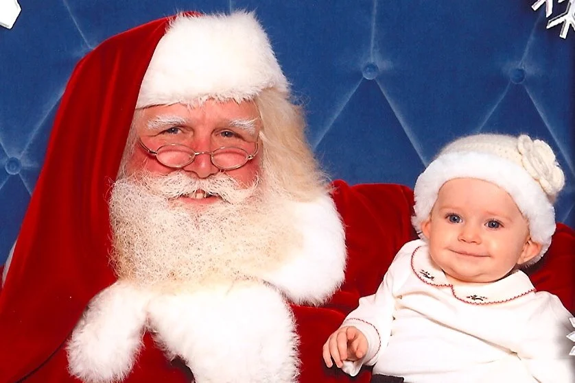 A man dressed as Santa Claus sitting with a smiling young girl wearing a white hat and holiday pajamas.