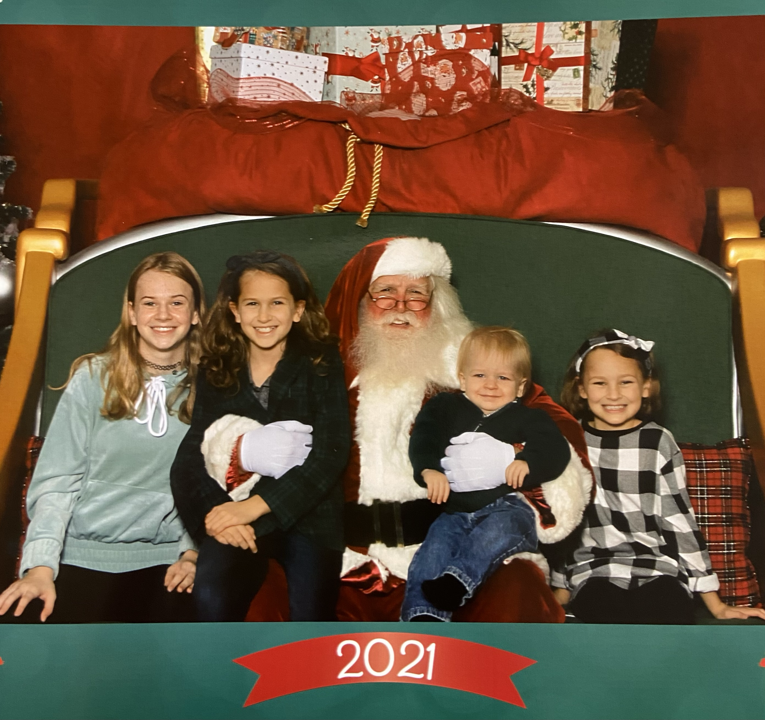 Children sitting with Santa Claus in a festive setting with Christmas gifts and decorations, 2021