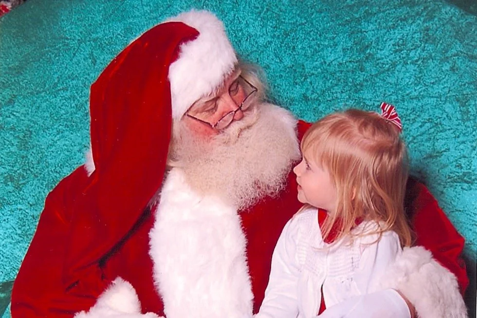 A young girl sitting on Santa Claus's lap, looking at him and smiling. Santa is dressed in a traditional red suit with a white beard and glasses. The background is teal.
