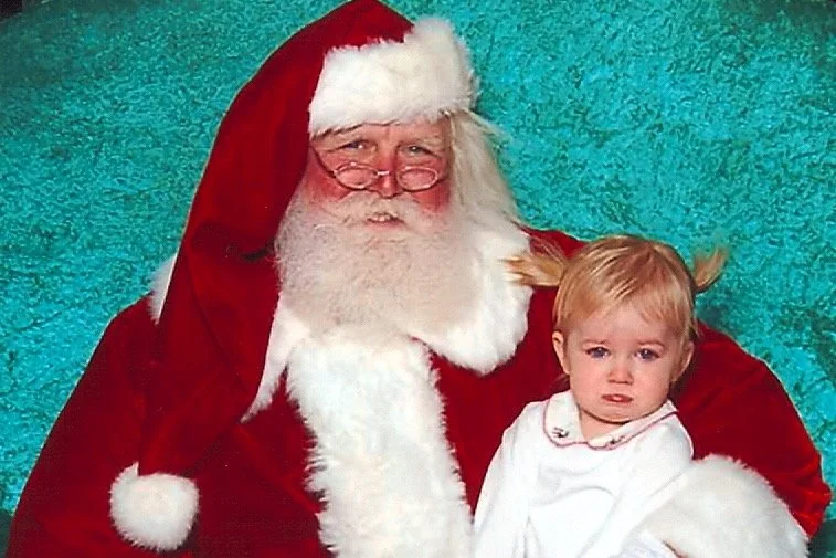 A person dressed as Santa Claus with glasses and a white beard, sitting next to a young girl with blonde hair tied in pigtails, against a teal background.