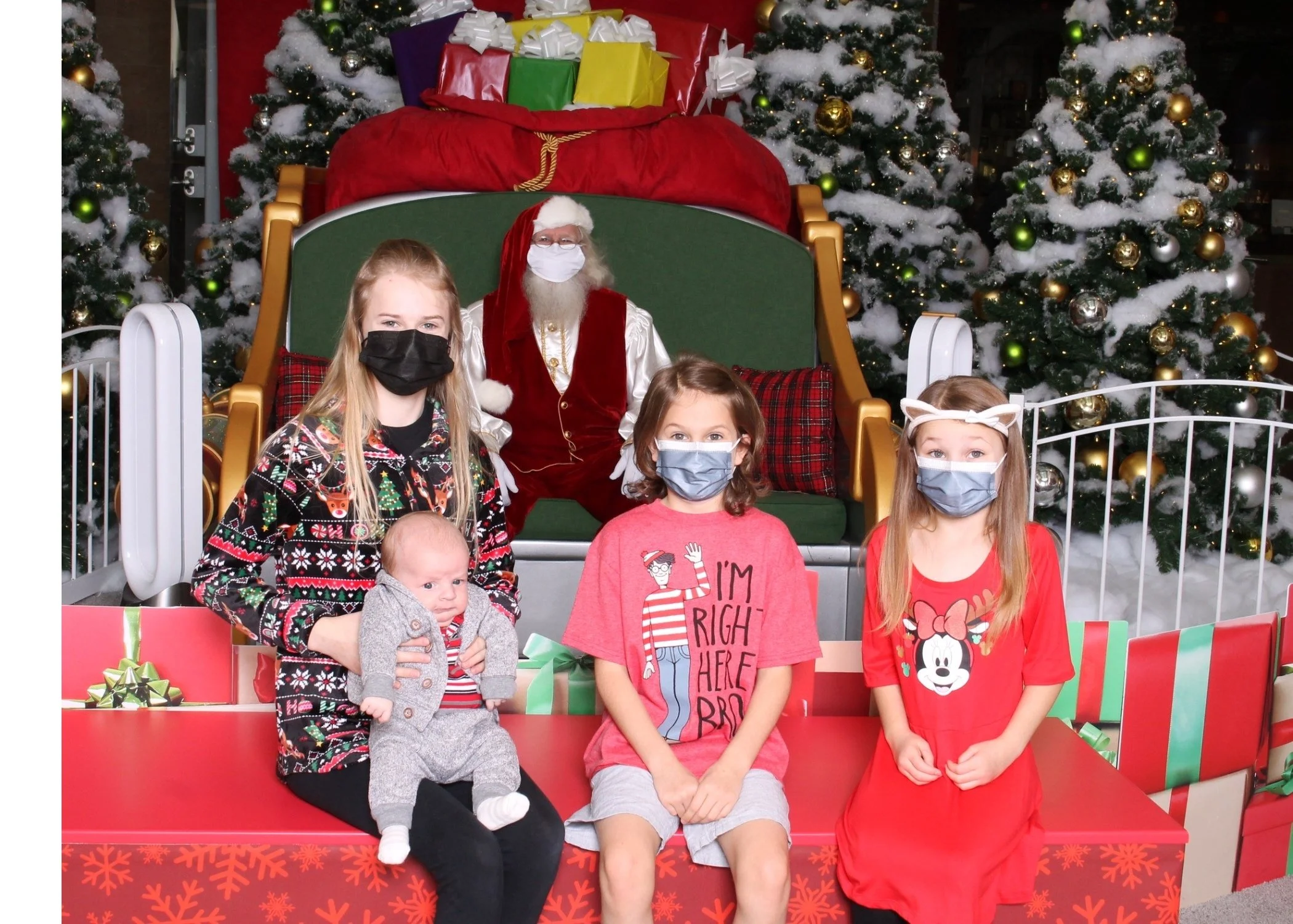 Group of children sitting in front of Santa Claus who is sitting on a large green throne, with decorated Christmas trees and wrapped presents in the background. All children and Santa are wearing face masks.