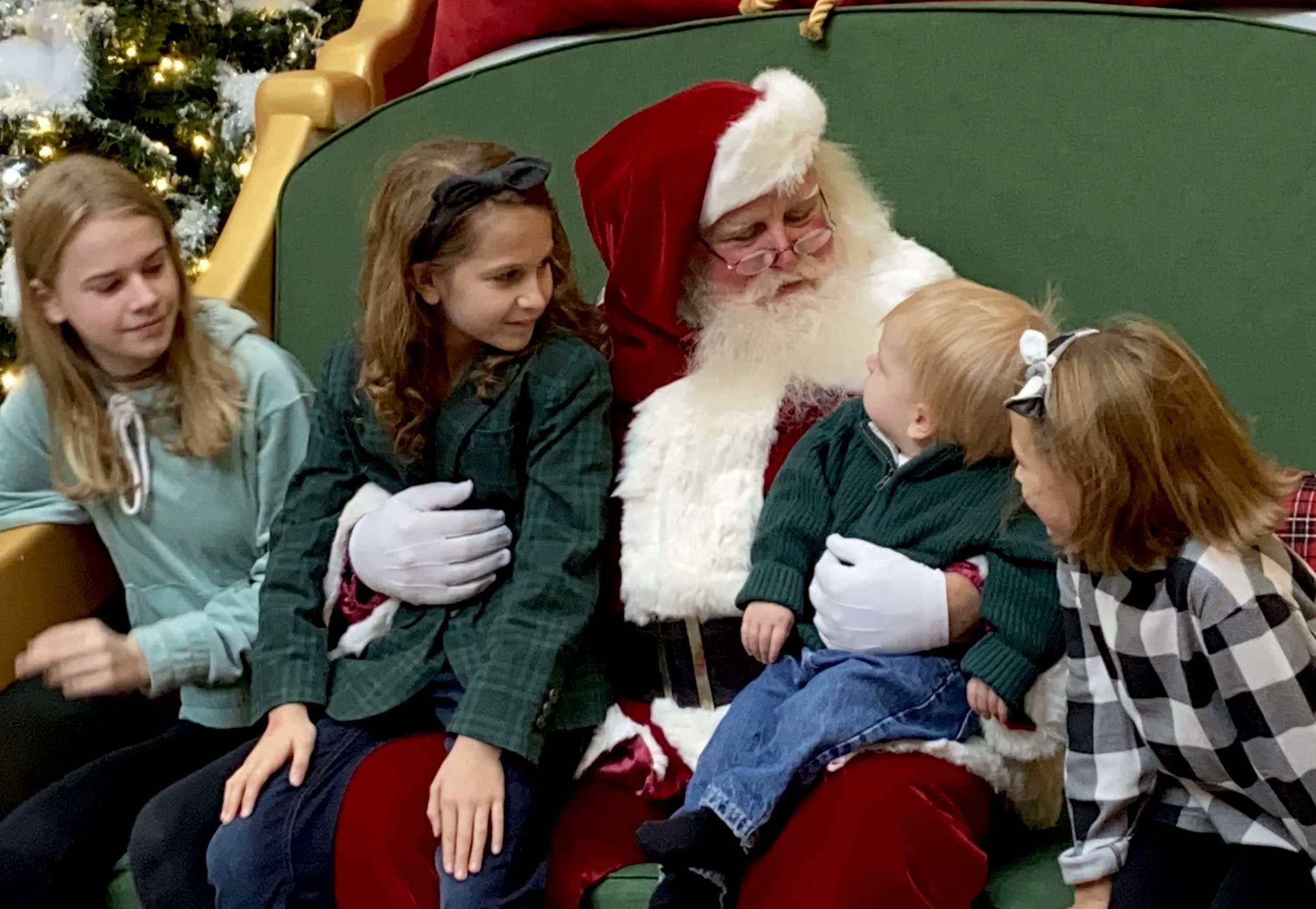 Santa Claus sitting on a green bench, talking to three young children during a Christmas event, with Christmas trees decorated with lights and ornaments in the background.