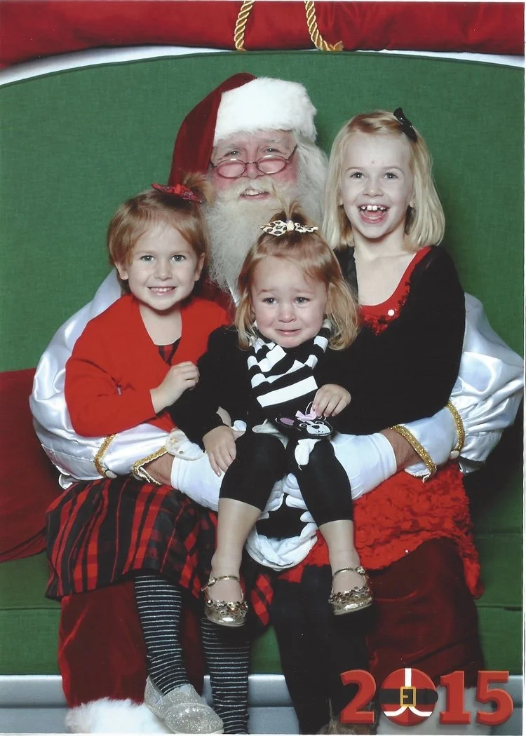 Three young girls sitting with Santa Claus on a green and red holiday backdrop, all smiling. The girls are dressed in festive holiday attire, and Santa wears his traditional red hat and suit. The year 2015 is displayed in red at the bottom right corn