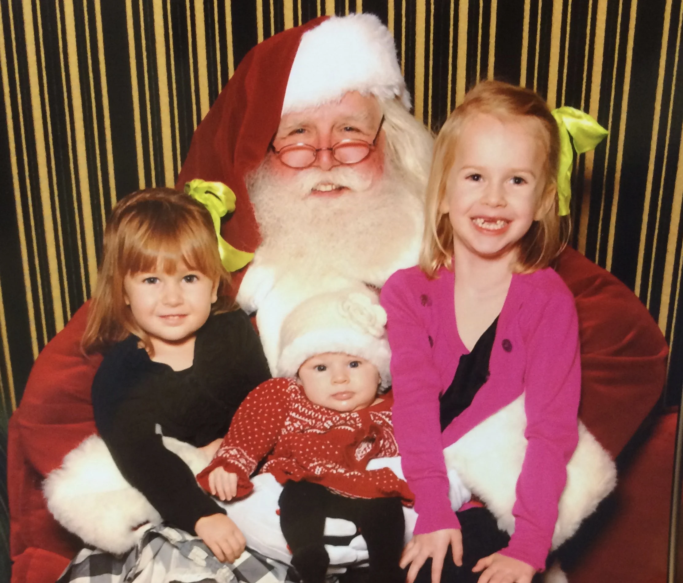 Four children sitting on Santa Claus's lap, who is wearing his traditional red suit and hat, with a black and gold striped curtain in the background.