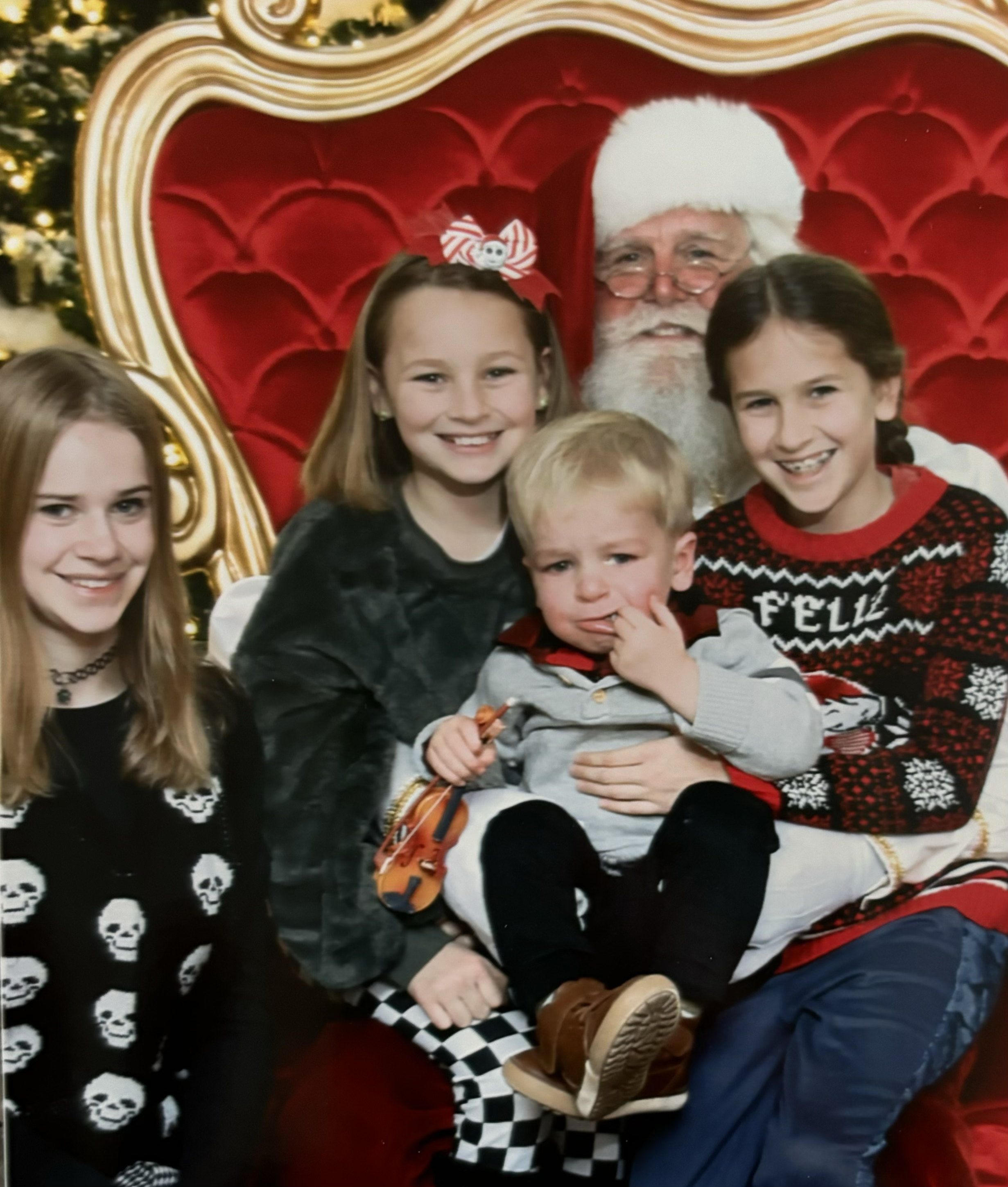 Group photo with Santa Claus on a red velvet throne, surrounded by children, in front of a Christmas decorated background.