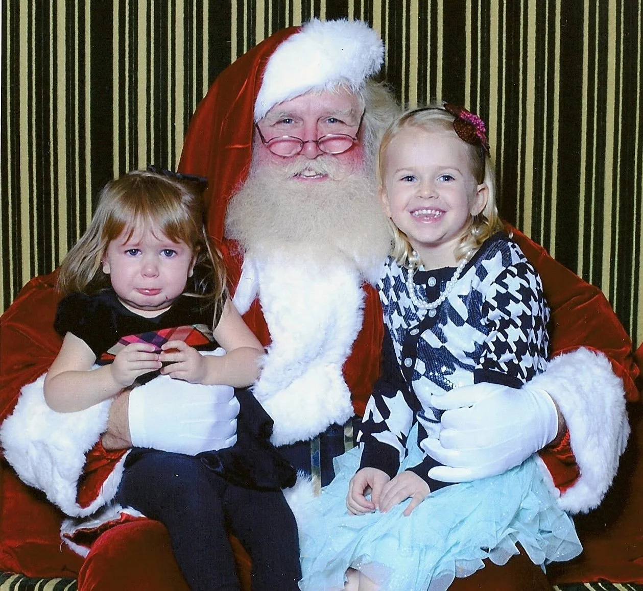 A person dressed as Santa Claus sitting on a red velvet chair with two young girls, one crying and the other smiling, against a black and gold striped curtain background.