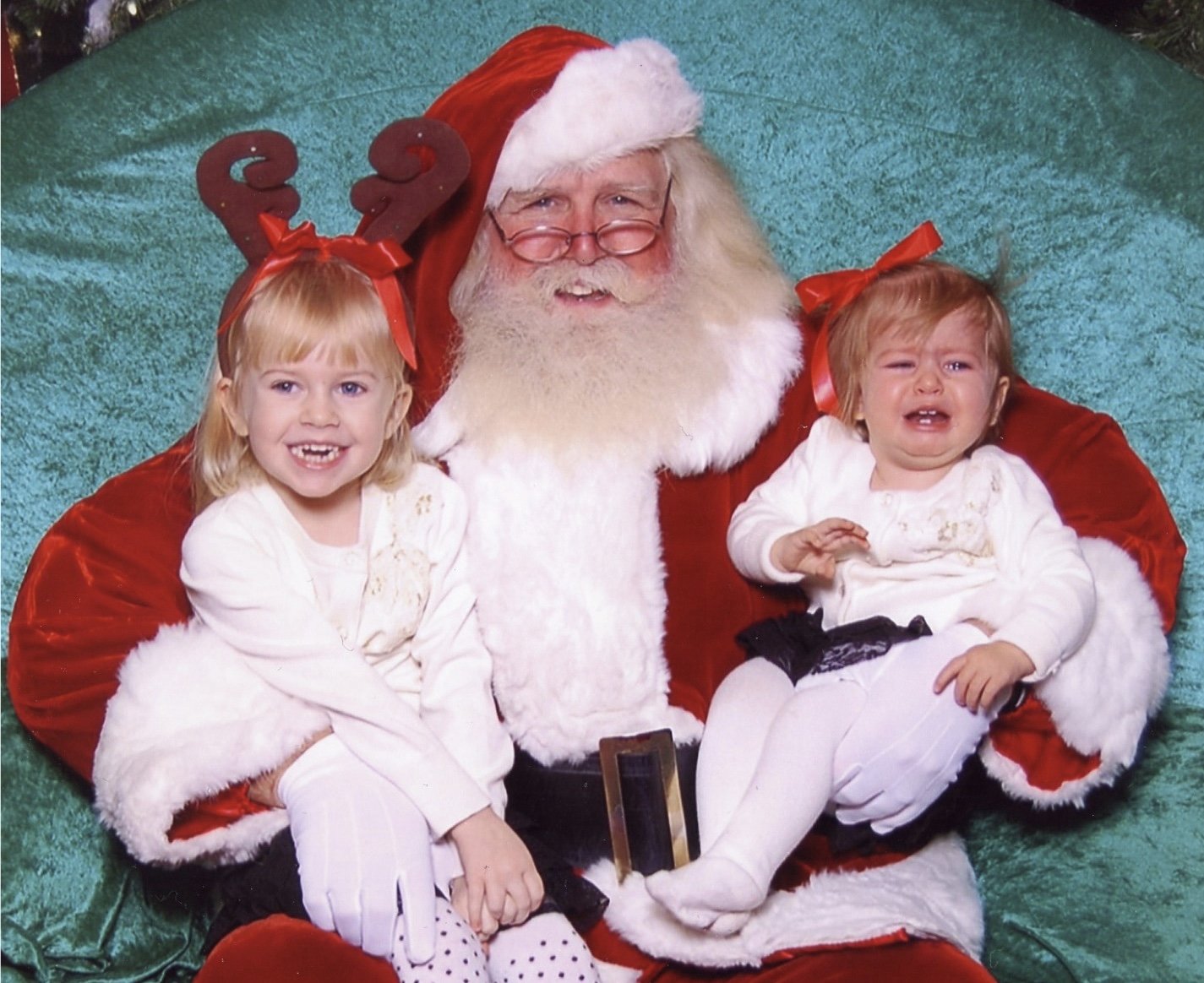Two young girls sitting on Santa Claus's lap during a Christmas photo. One girl is smiling happily, and the other girl appears to be crying or upset. Santa is wearing a traditional red suit with white fur trim, glasses, and a red hat, while the girls