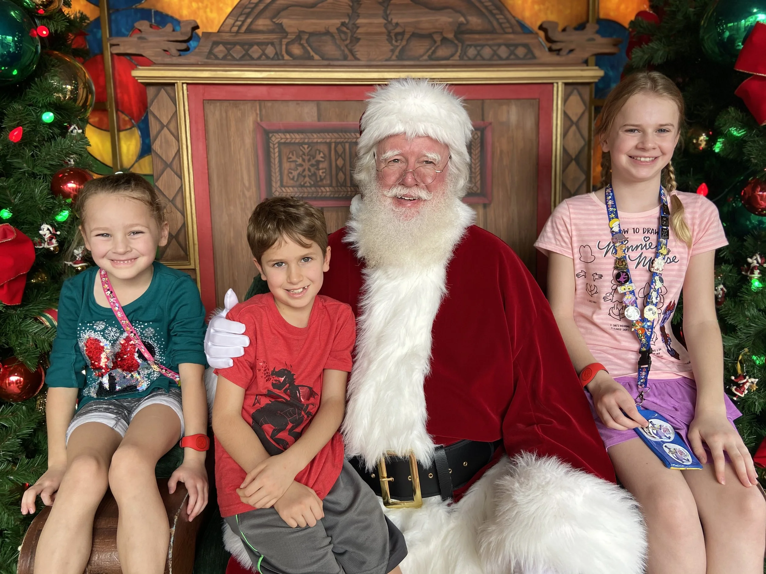 Four children sitting with Santa Claus during Christmas, surrounded by decorated Christmas trees with ornaments and lights.