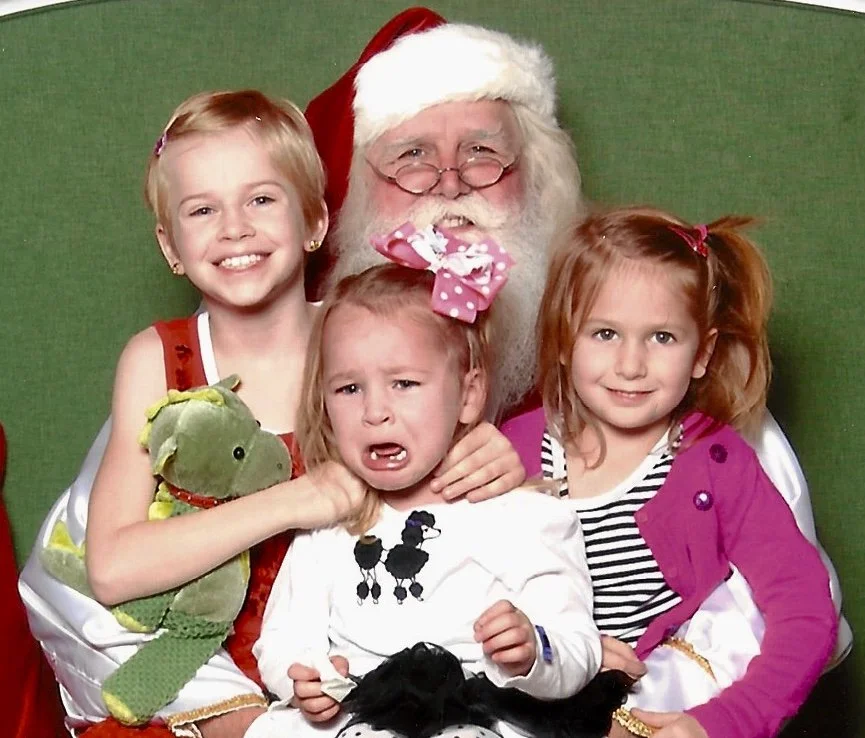 Three young girls and Santa Claus posing for a holiday photo, with one girl crying and holding a green stuffed toy, in front of a green backdrop.