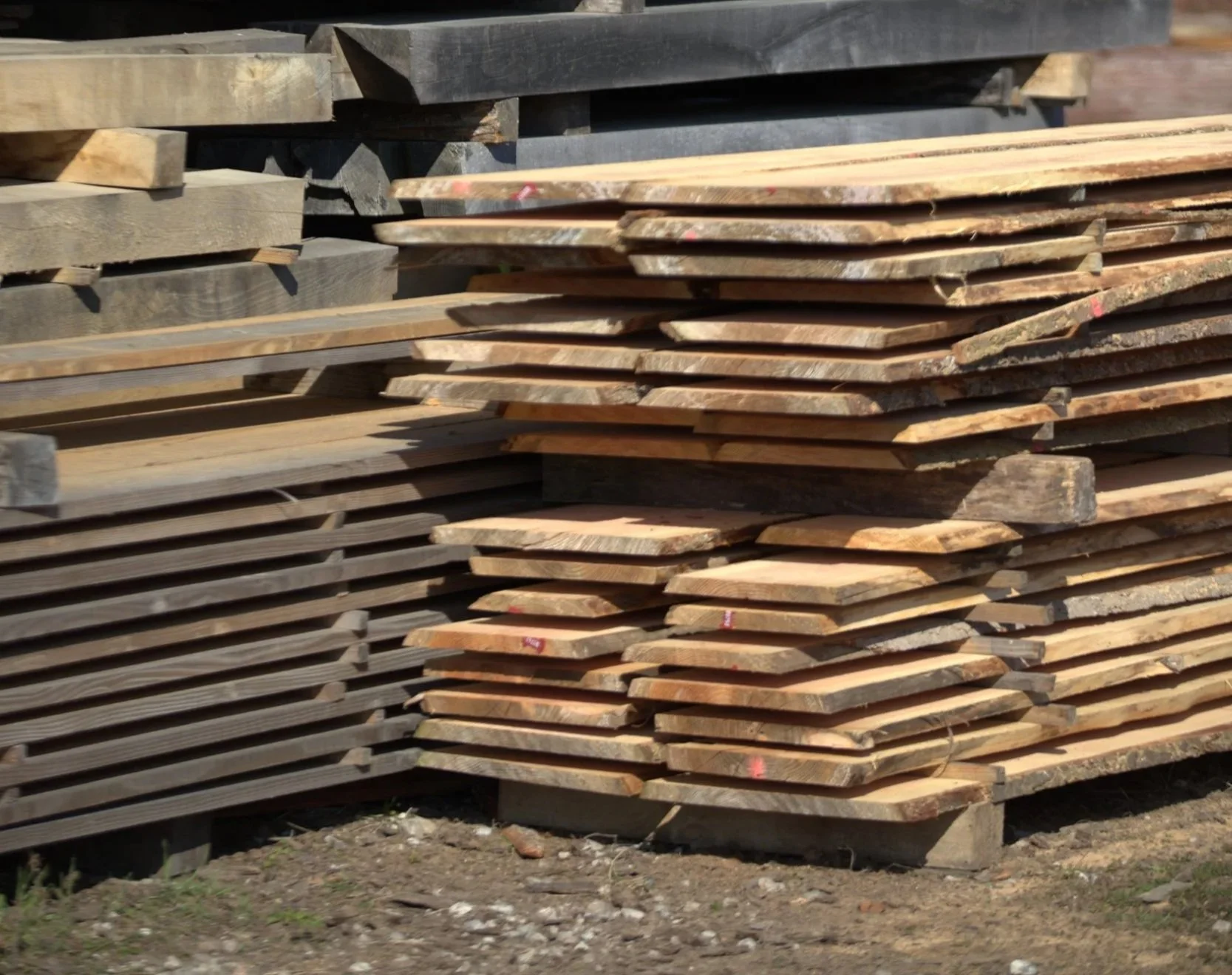 Stack of cut lumber and wooden planks outdoors on a work surface with a house and bushes in the background
