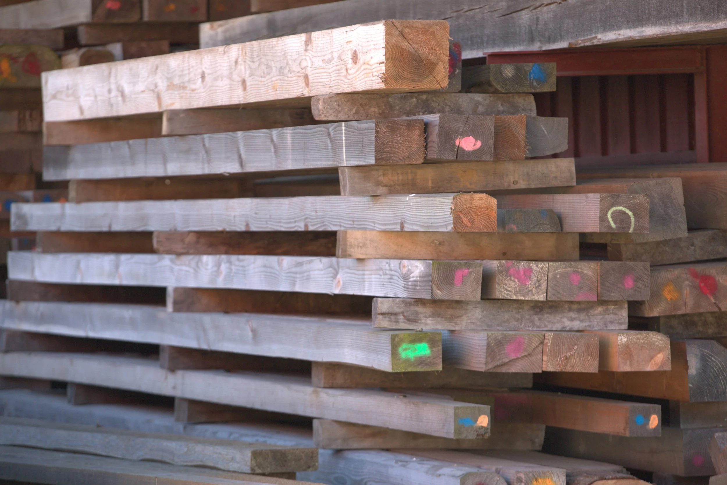 Stacked lumber outdoors against a shelter, with snow on the ground and woodworking marks on some pieces.