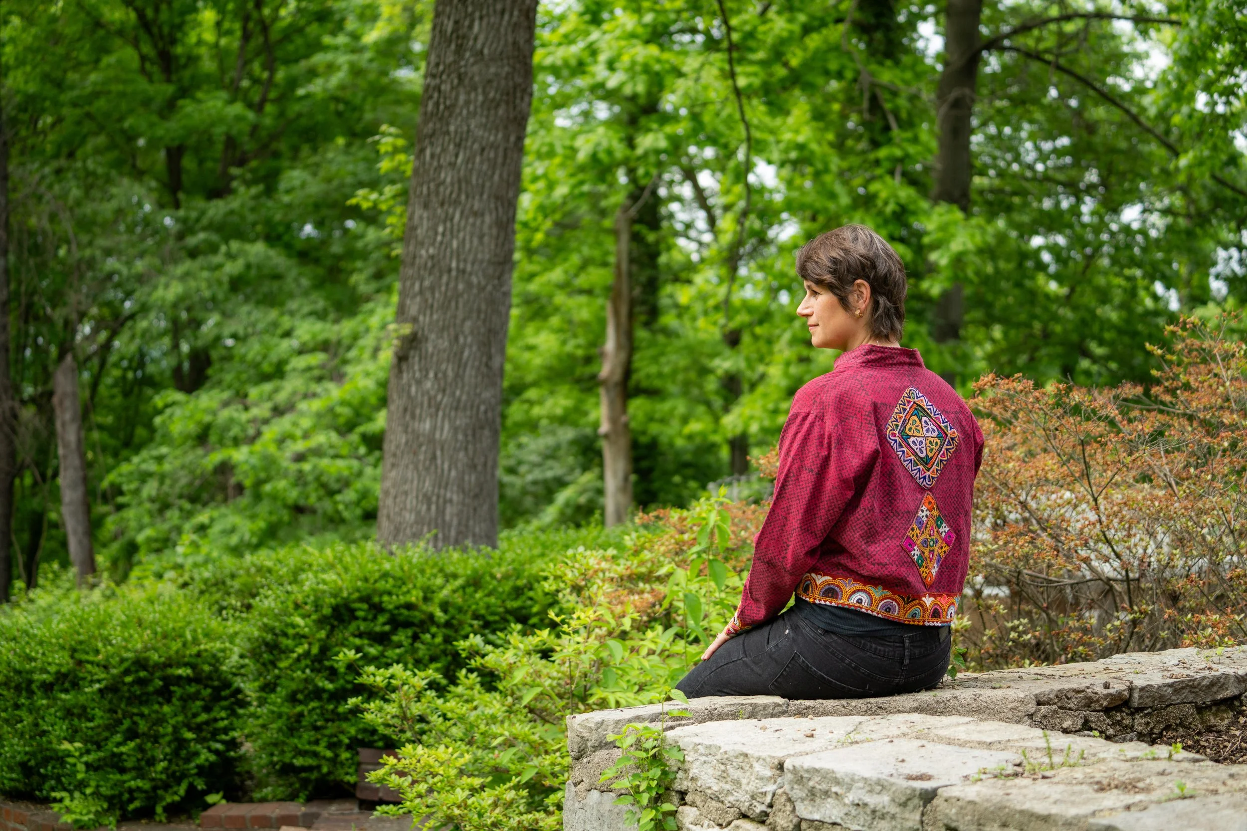 A woman sitting on a stone ledge in a lush green park, wearing a red patterned jacket and black pants, looking to the side.