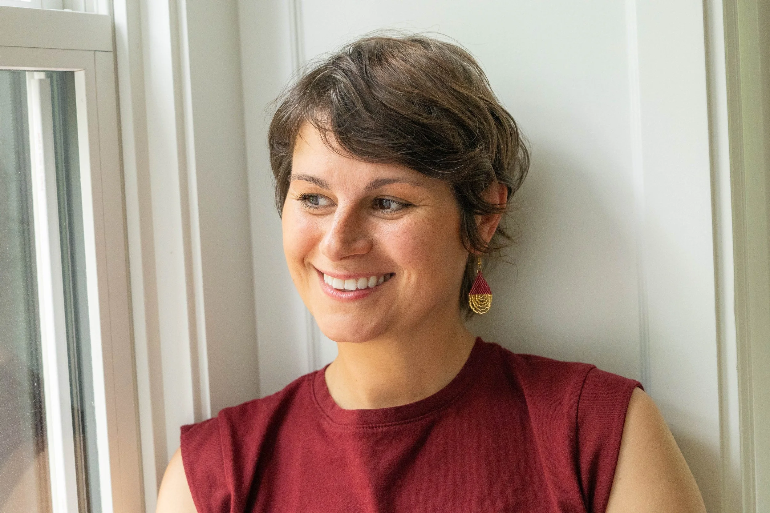 A woman with short brown hair and light skin, smiling, wearing a sleeveless burgundy top and gold and pink earrings, standing near a window with white paneling.