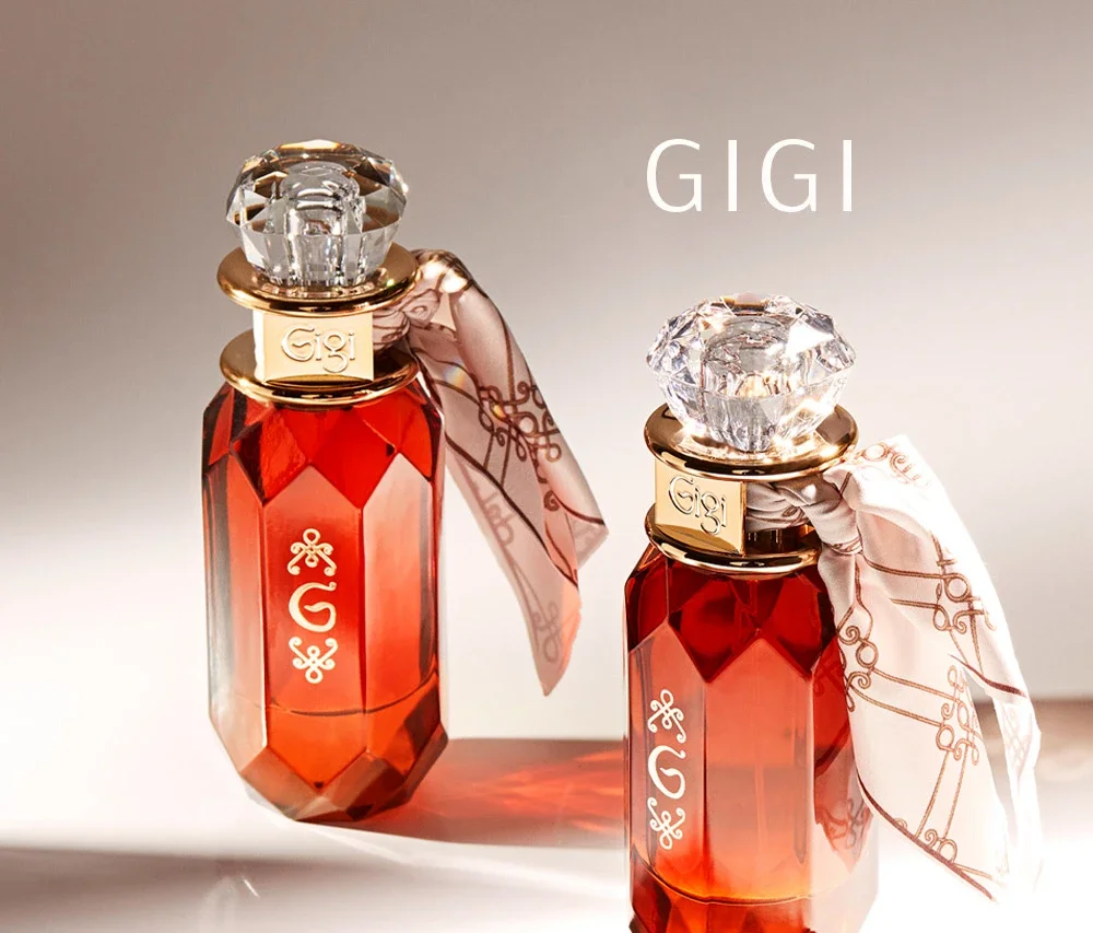 Dois frascos de perfume com tampas de cristal e detalhes dourados, um com lenço branco decorativo, sobre fundo claro e com a palavra GIGI escrita ao lado