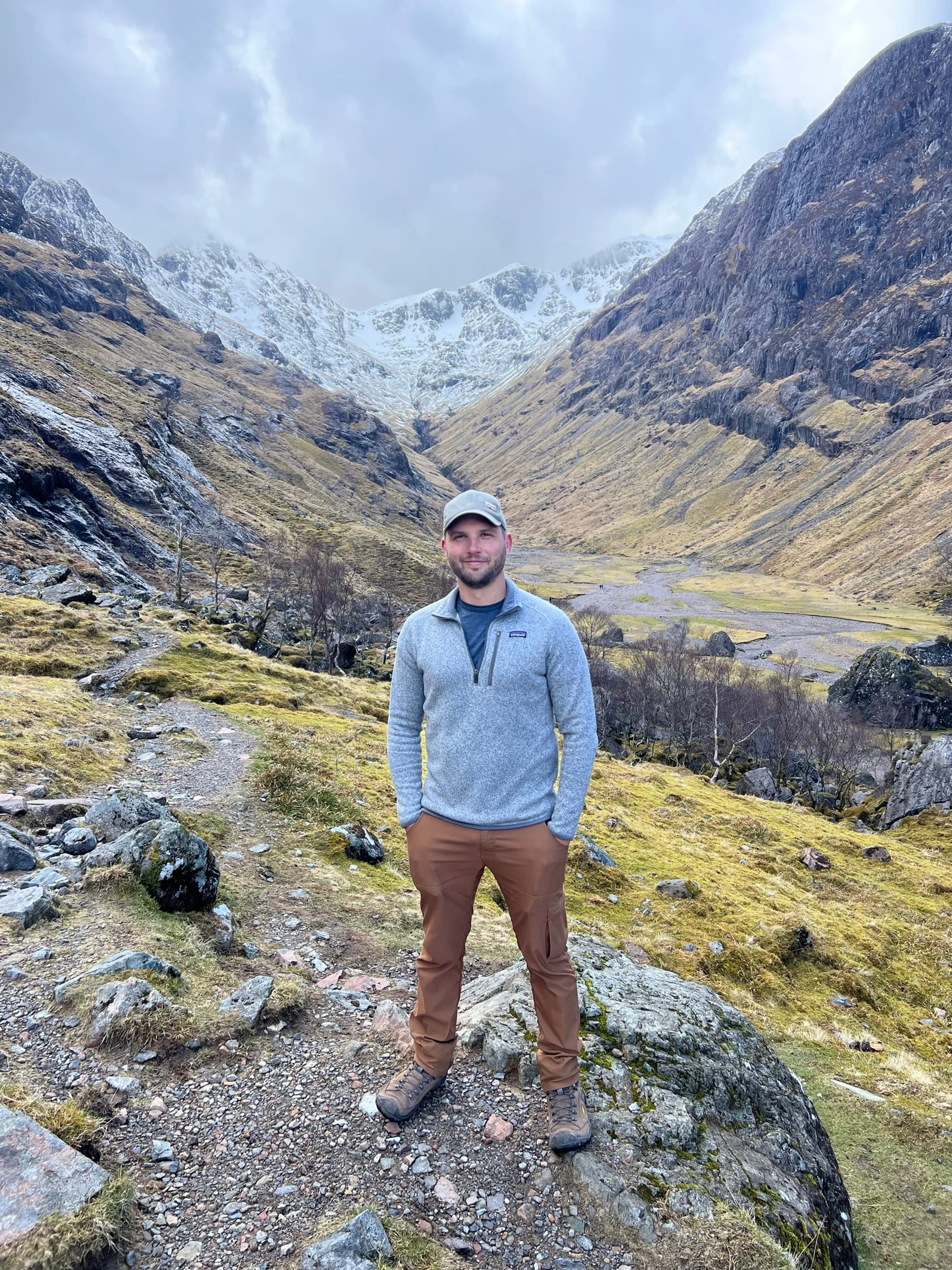 A man in outdoor hiking gear standing on a rocky trail in a mountainous landscape with snow-capped peaks and grassy slopes.