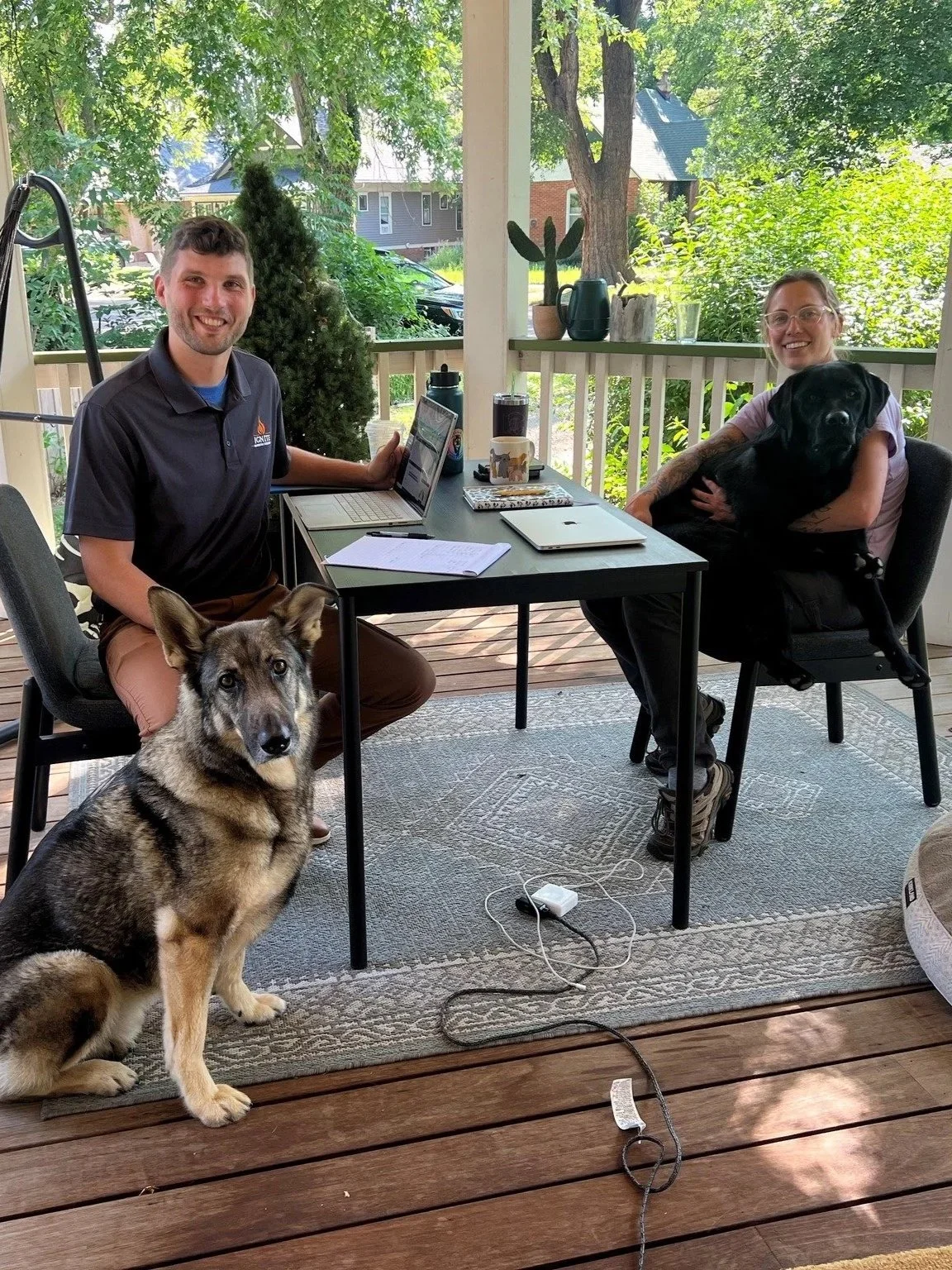 Two people sitting at a table with a dog in each arm, on a porch with trees and houses visible in the background. The person on the left is smiling, the person on the right is holding a large black dog, and a dog is sitting on the floor.