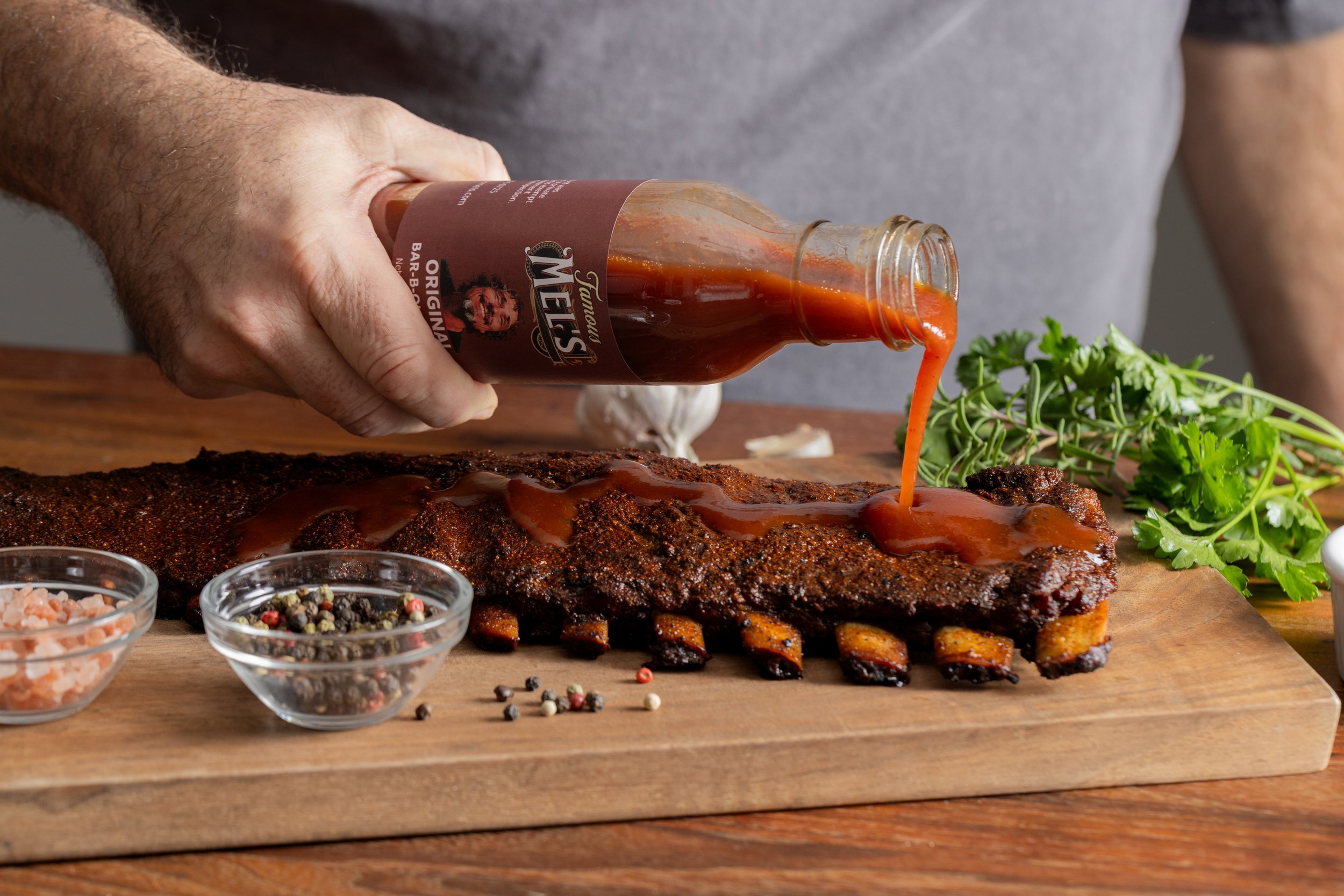 Man pouring barbecue sauce on a rack of cooked ribs on a wooden cutting board with herbs and seasoning in bowls.