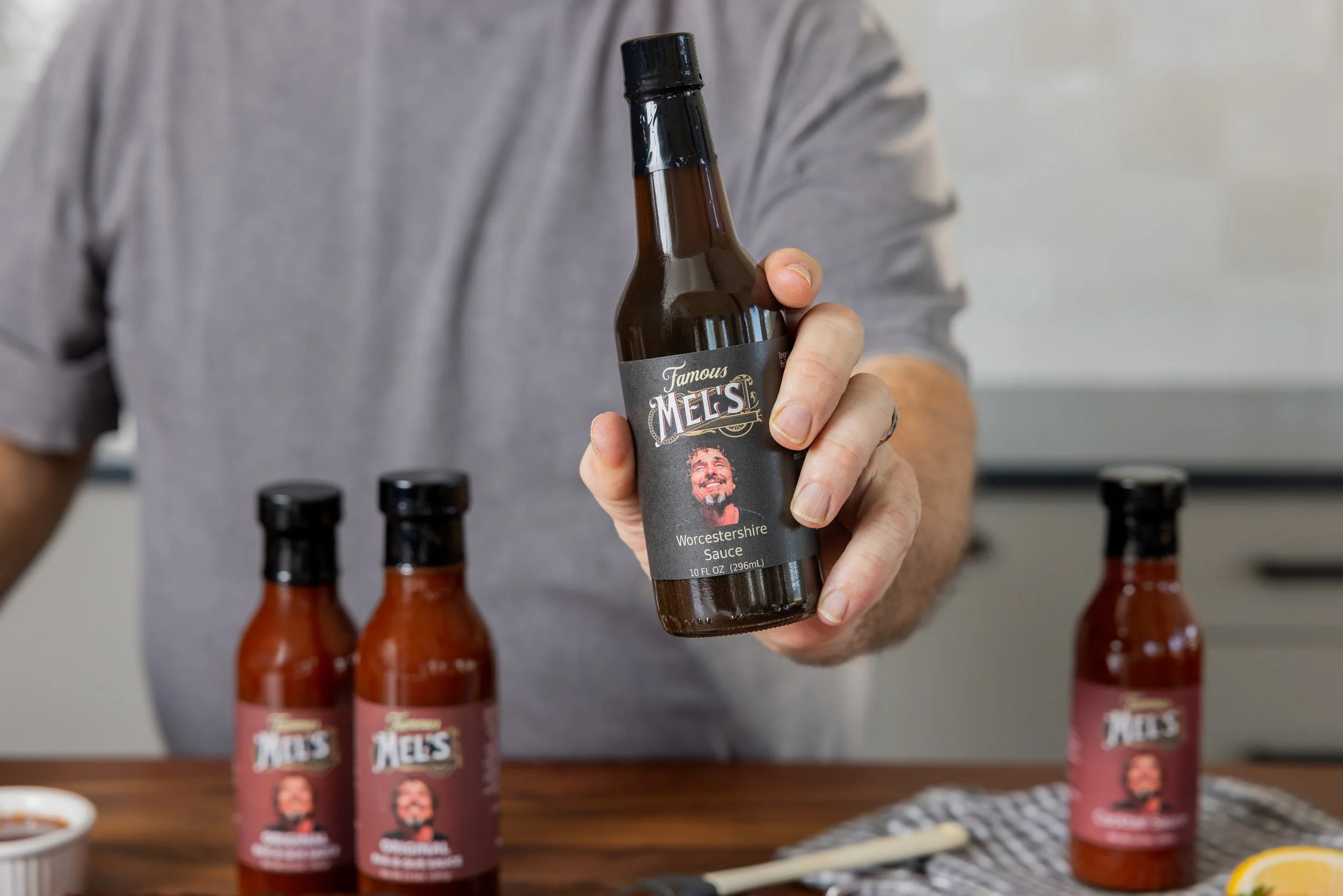 Person holding a bottle of Mel's Worcestershire Sauce with three other bottles on a wooden table.