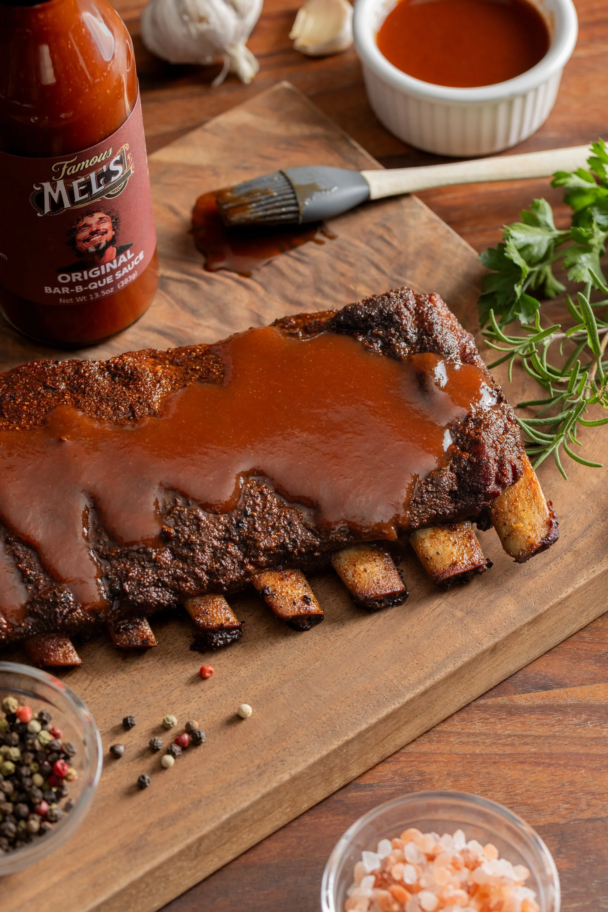 A rack of BBQ ribs with sauce on a wooden cutting board, surrounded by garlic, herbs, pepper, and salt, with barbecue sauce bottle and bowls of sauce on a wooden table.