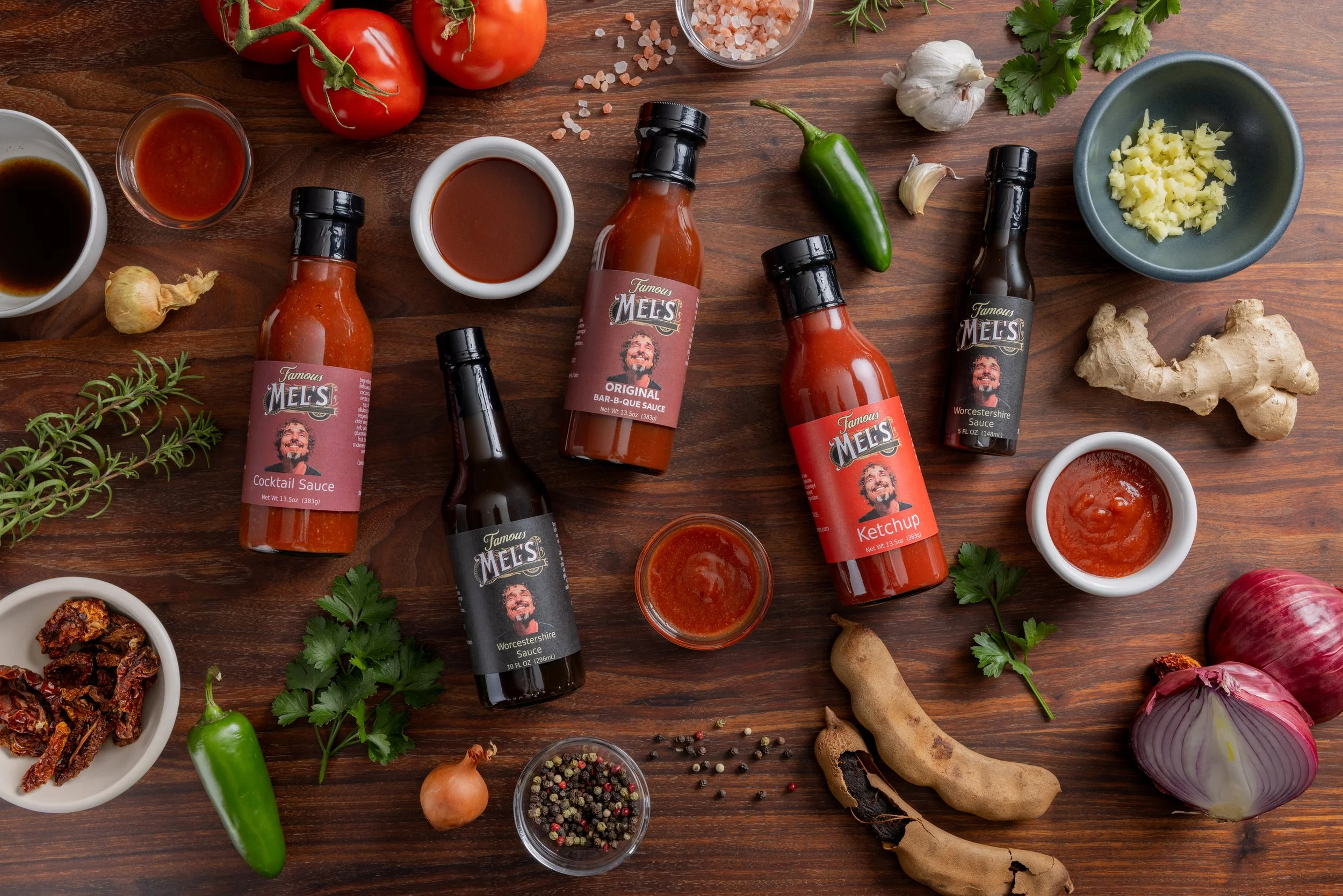 Assorted bottles of barbecue sauces, fresh vegetables including tomatoes, garlic, ginger, onions, chili peppers, and herbs on a wooden surface, along with small bowls of sauces, dried chili peppers, peppercorns, and salt.
