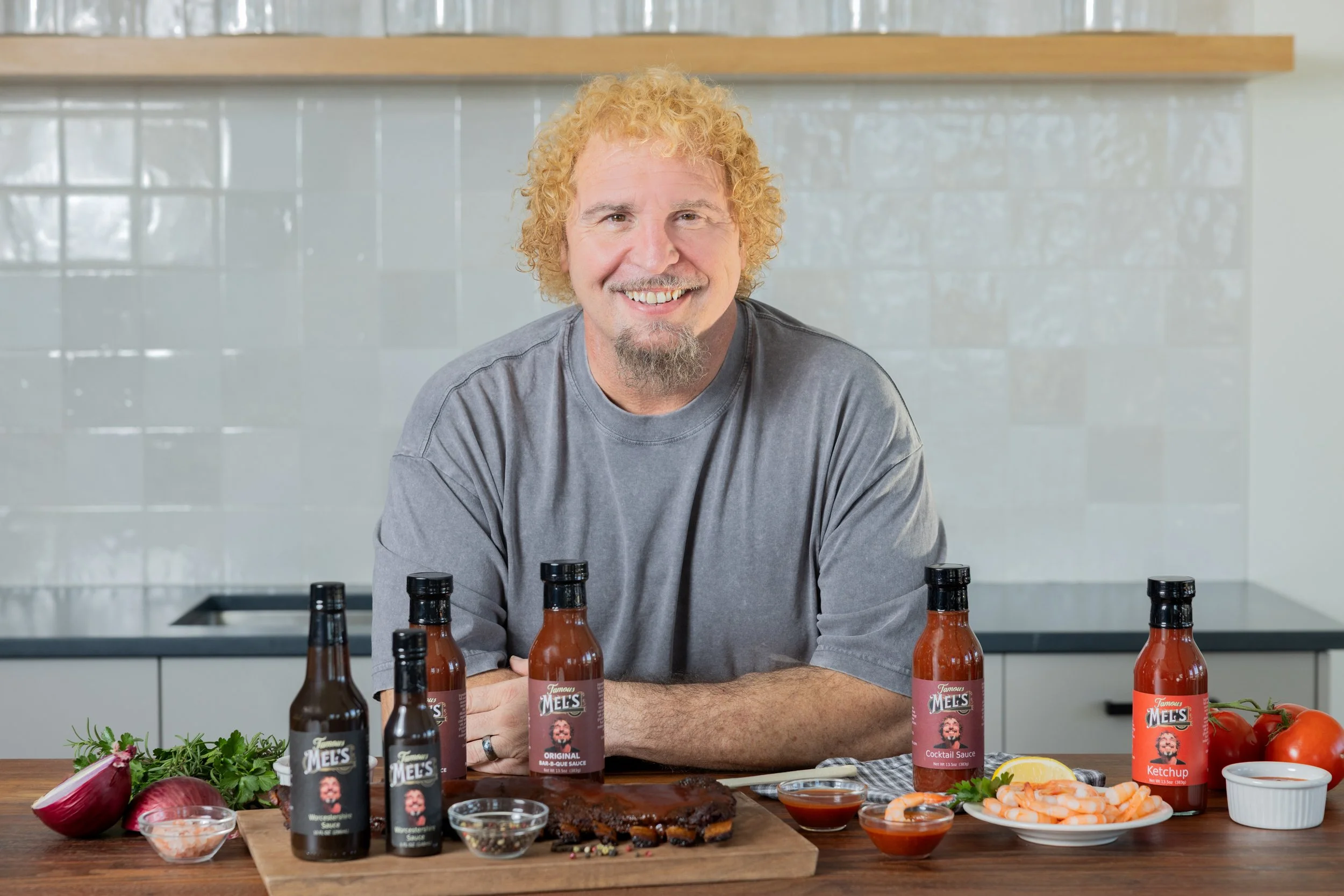 A man with curly blond hair and a goatee smiling at the camera, seated at a kitchen counter with bottles of barbecue sauces, a plate of cooked ribs, garnishes, and condiments for preparing barbecue.