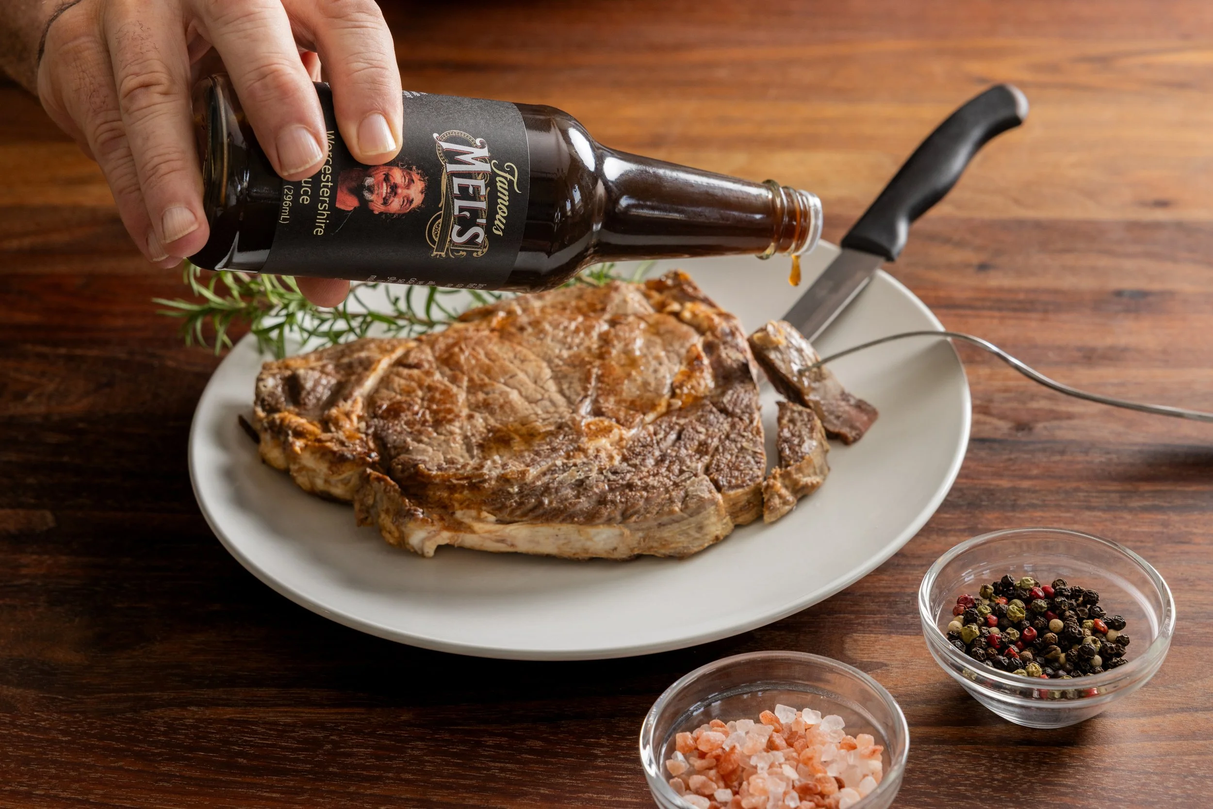 Someone pouring Worcestershire sauce onto a grilled T-bone steak on a white plate, with black pepper and pink Himalayan salt in small bowls nearby, on a wooden table.